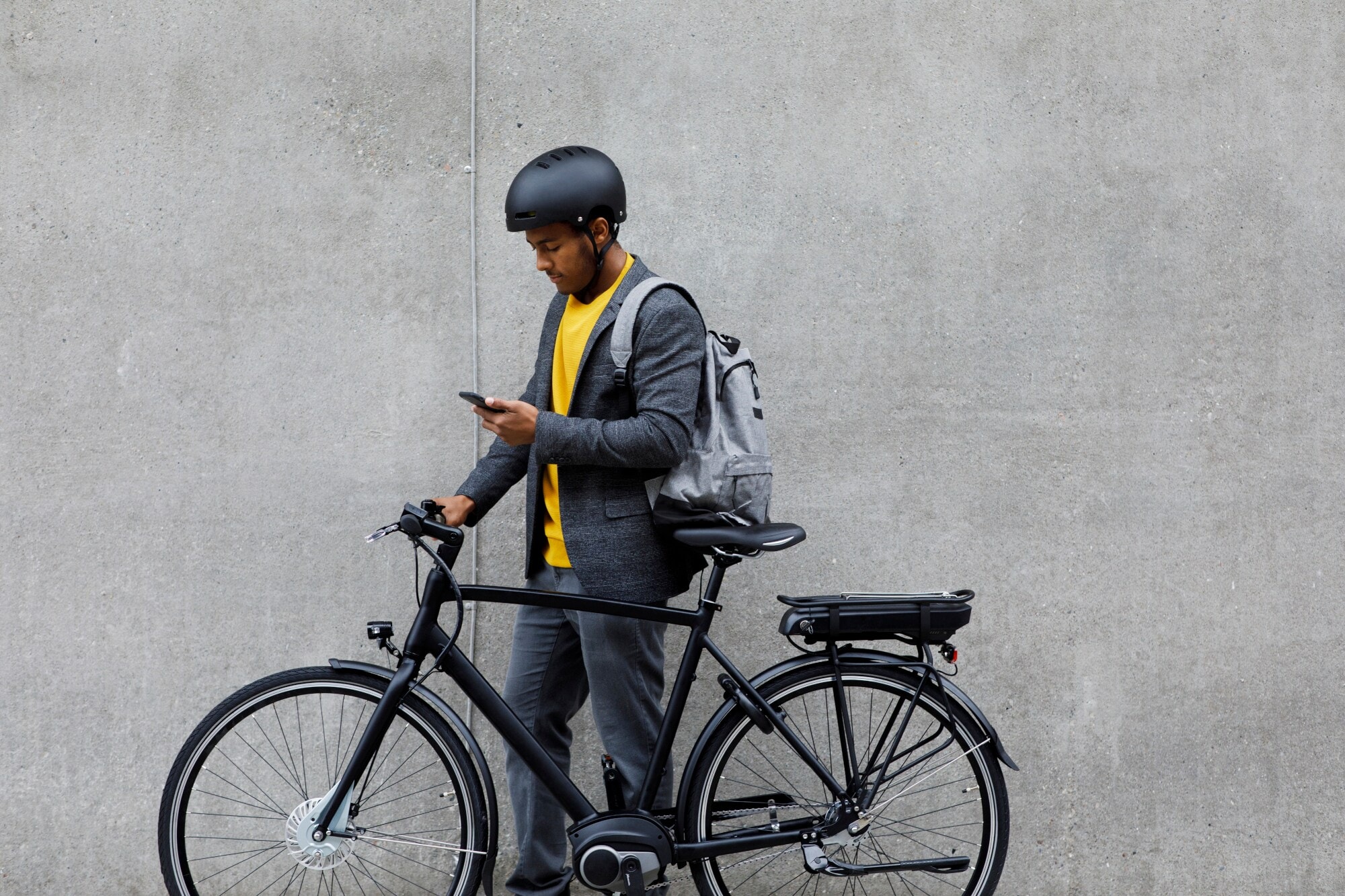 Ein junger Mann mit Rucksack und Fahrradhelm steht neben seinem E-Bike und schaut auf sein Smartphone. Ein junger Mann mit Rucksack und Fahrradhelm steht neben seinem E-Bike und schaut auf sein Smartphone.