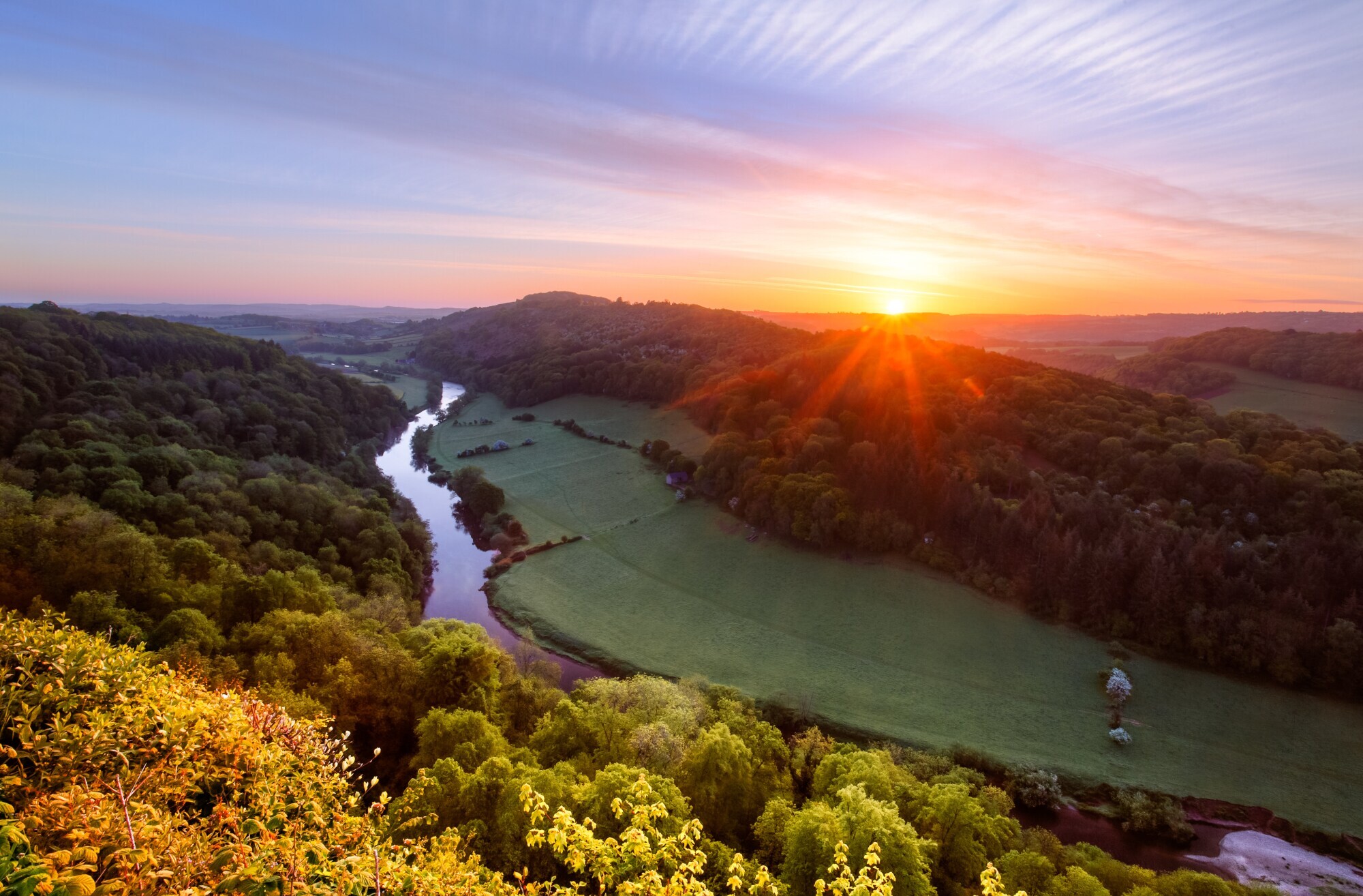 Blick auf ein grünes Flusstal, umgeben von Waldlandschaft, bei Sonnenaufgang. Blick auf ein grünes Flusstal, umgeben von Waldlandschaft, bei Sonnenaufgang.