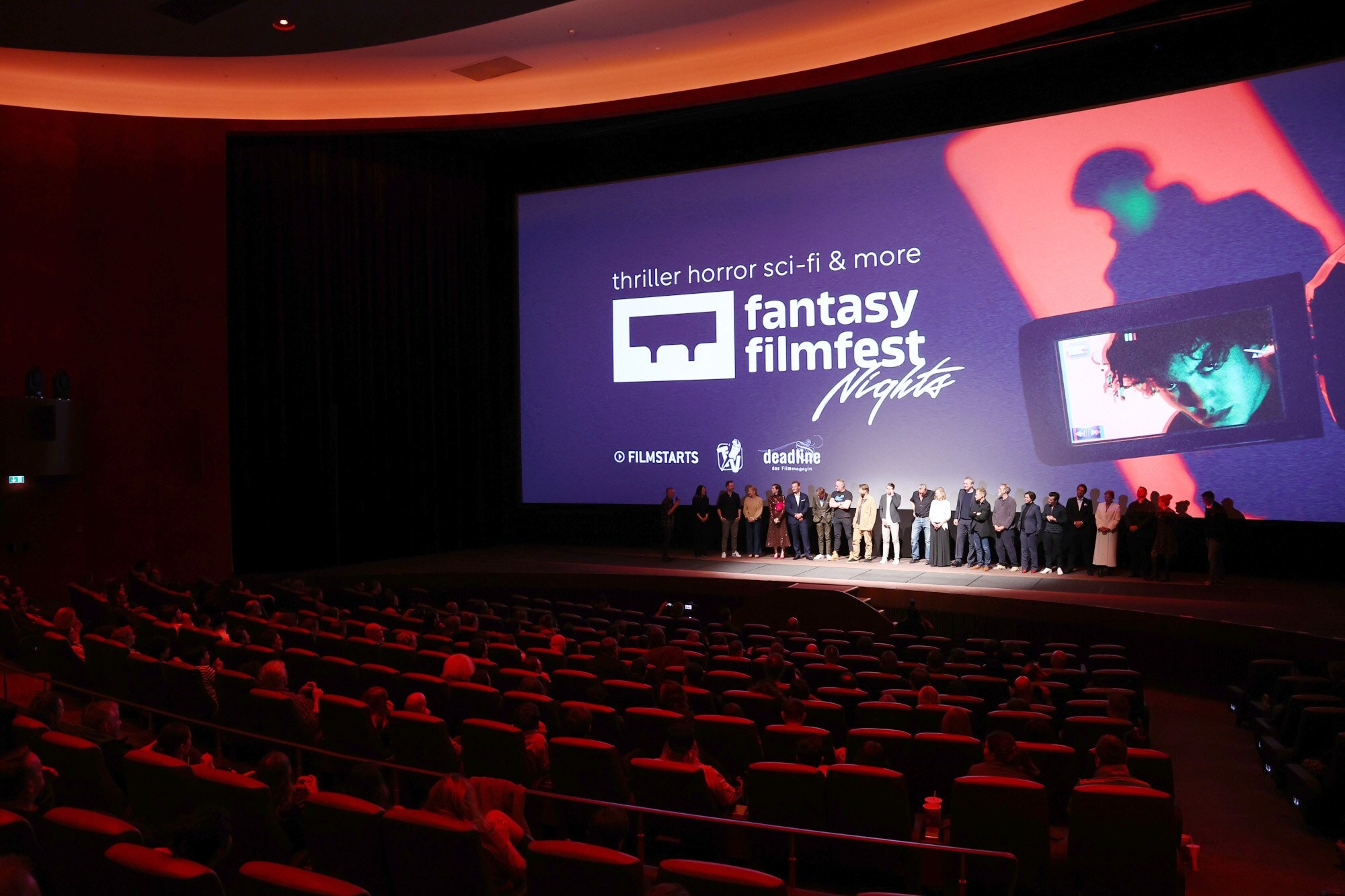 Eine Personengruppe steht vor einer Leinwand auf einer Bühne vor Publikum in roten Samtsesseln in einem Kinosaal Eine Personengruppe steht vor einer Leinwand auf einer Bühne vor Publikum in roten Samtsesseln in einem Kinosaal.