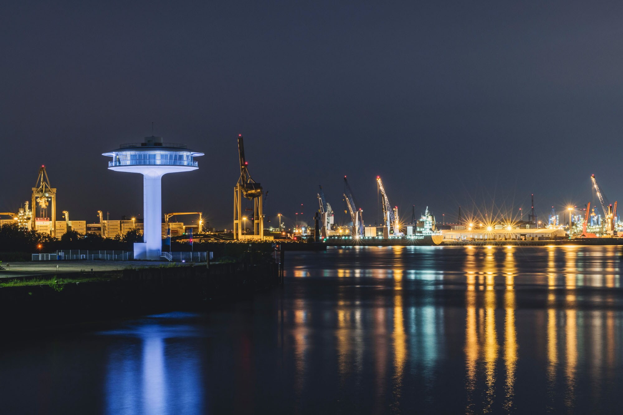 Beleuchteter Leuchtturm in Ufo-Form mit Panoramafenstern vor der Skyline des beleuchteten Hamburger Hafens bei Nacht.
