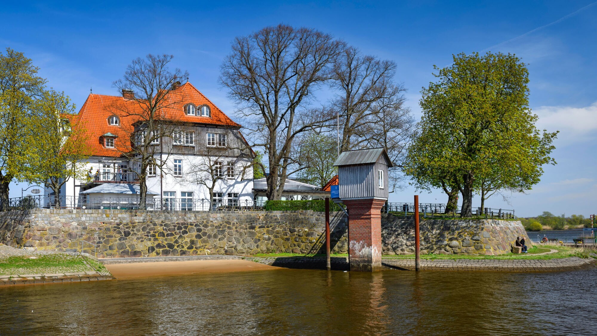 Ein Pegelhaus im Wasser vor einer Uferpromenade mit Fährhaus in begrünter Umgebung.