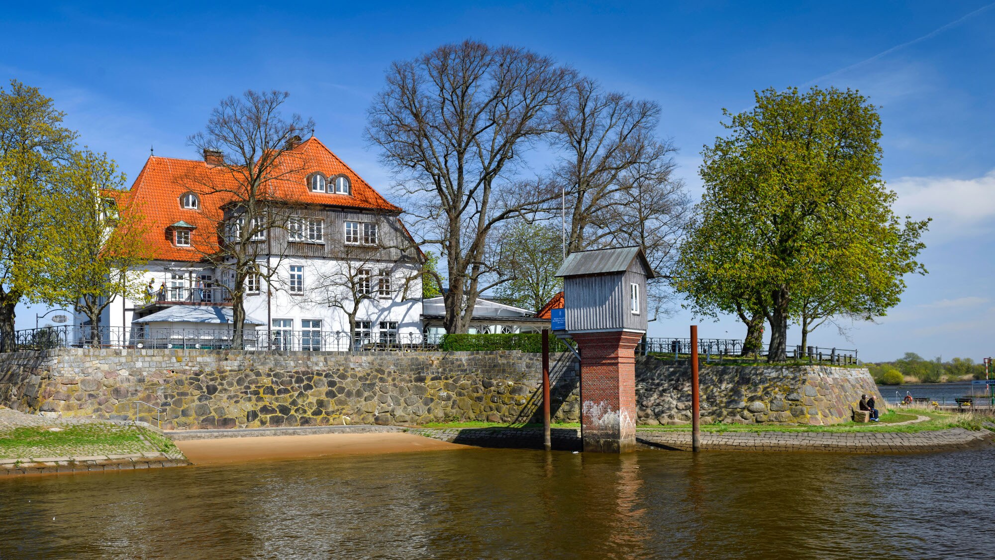 Ein Pegelhaus im Wasser vor einer Uferpromenade mit Fährhaus in begrünter Umgebung. Ein Pegelhaus im Wasser vor einer Uferpromenade mit Fährhaus in begrünter Umgebung.