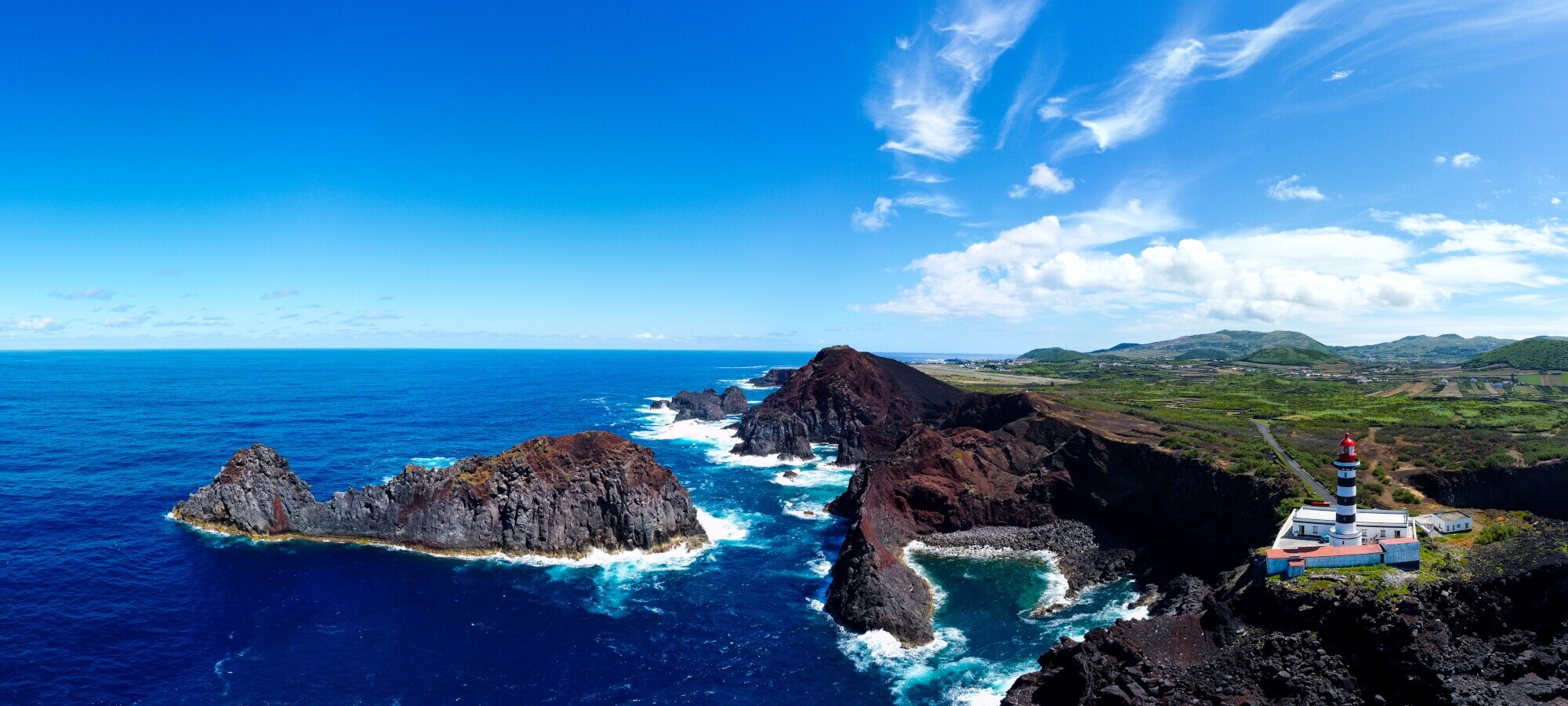 Luftaufnahme einer schroffen Felsküste mit Leuchtturm auf einer Klippe und einer vorgelagerter Felseninsel in Walfischform.