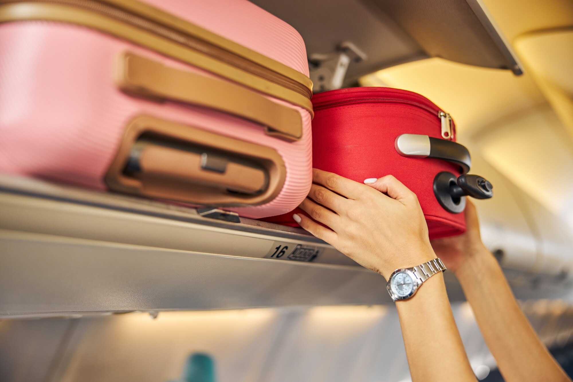 Zwei Hände greifen einen roten Koffer in einem Overhead-Bin eines Flugzeugs, im Vordergrund ein Koffer in Rosa. Zwei Hände greifen einen roten Koffer in einem Overhead-Bin eines Flugzeugs, im Vordergrund ein Koffer in Rosa.
