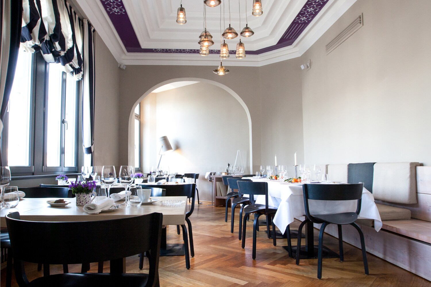 Heller Speisesaal eines edlen Restaurants mit schwarzen Holzstühlen an eingedeckten Tischen in einem klassischen Altbau mit hohen Decken und Holzparkett.