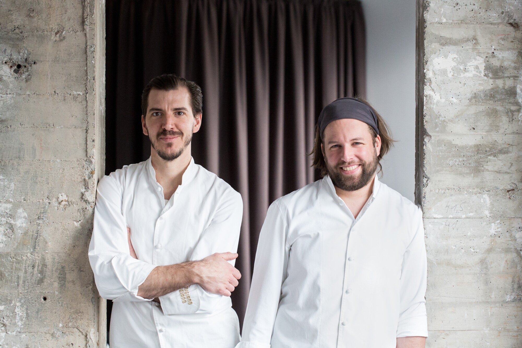 Porträts von zwei jungen Männern in weißen Kochjacken vor einer Betonwand mit Vorhang.