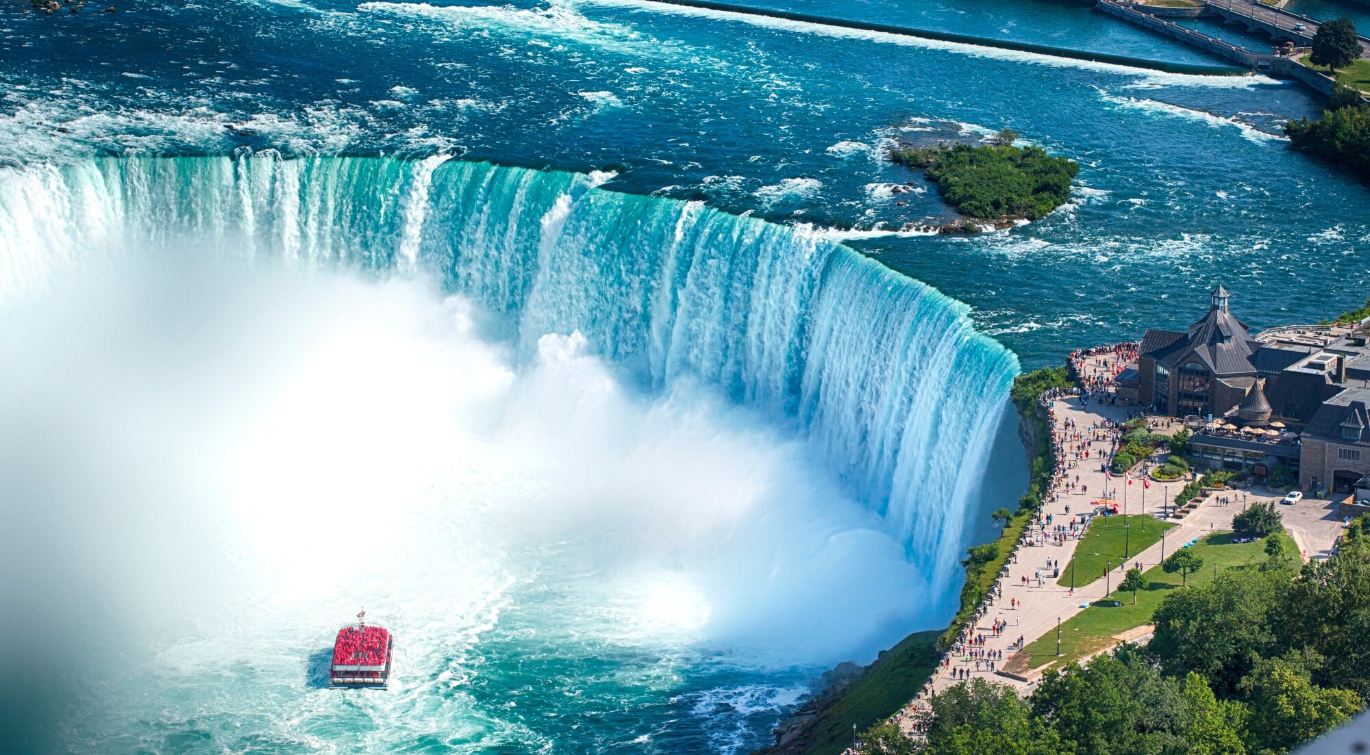 Luftaufnahme eines runden Wasserbeckens mit Boot vor einem Wasserfall, am Ufer eine Aussichtsplattform mit Personen. Luftaufnahme eines runden Wasserbeckens mit Boot vor einem Wasserfall, am Ufer eine Aussichtsplattform mit Personen.