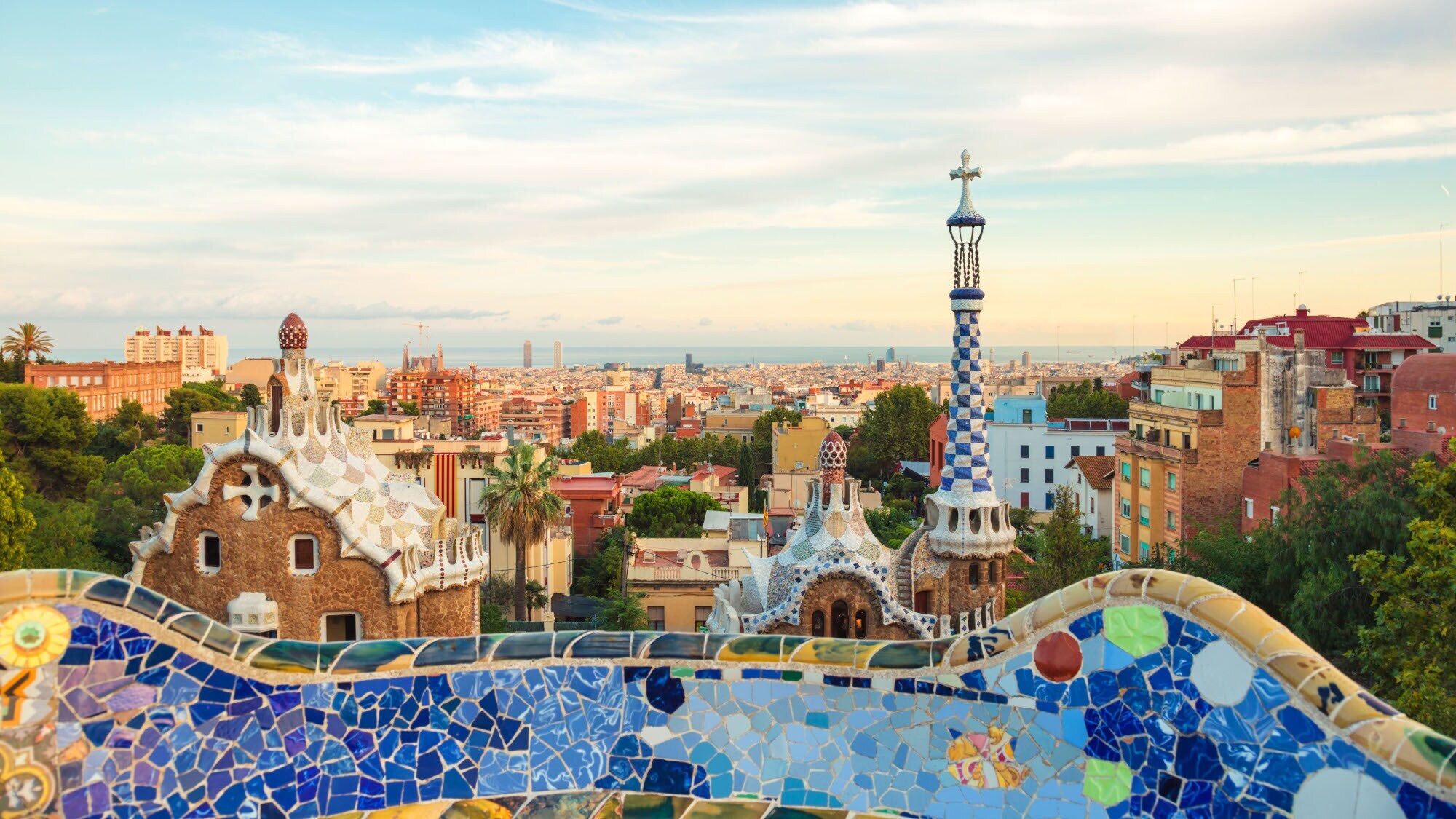kyline von Barcelona am Meer, im Vordergrund eine geschwungene Mauer mit Mosaiksteinen vor zwei weiteren Bauwerken von Gaudí.