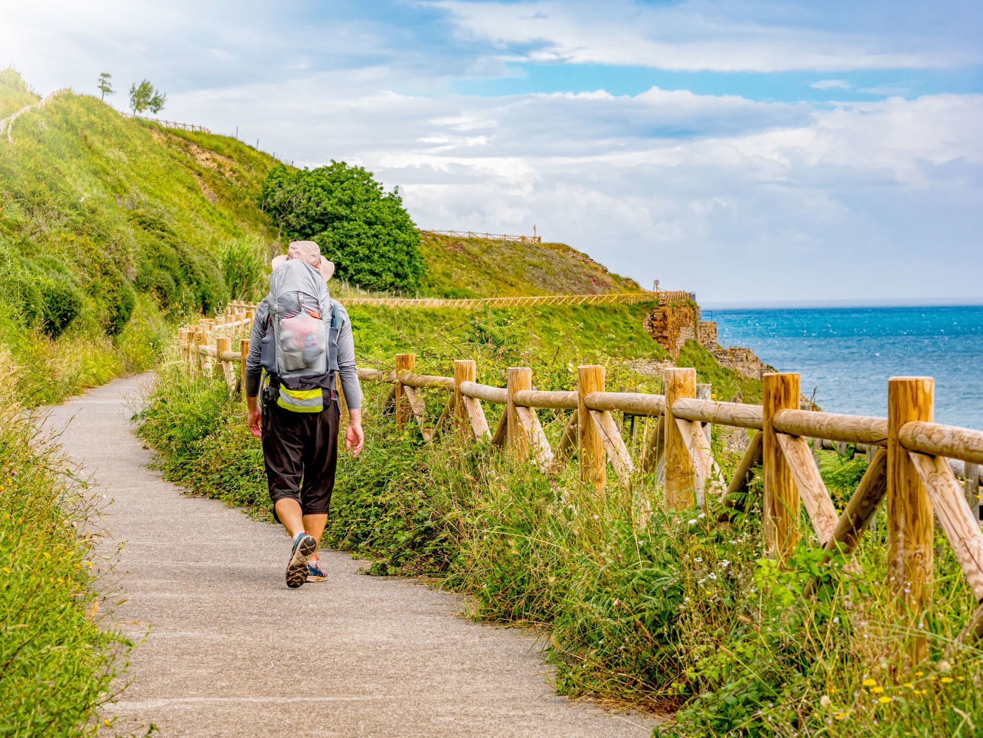 Lonely Pilgrim with backpack walking the Camino de Santiago in Spain, Way of St James Rückansicht eines Mannes mit Rucksack auf einem Wanderweg an einer begrünten Felsküste.