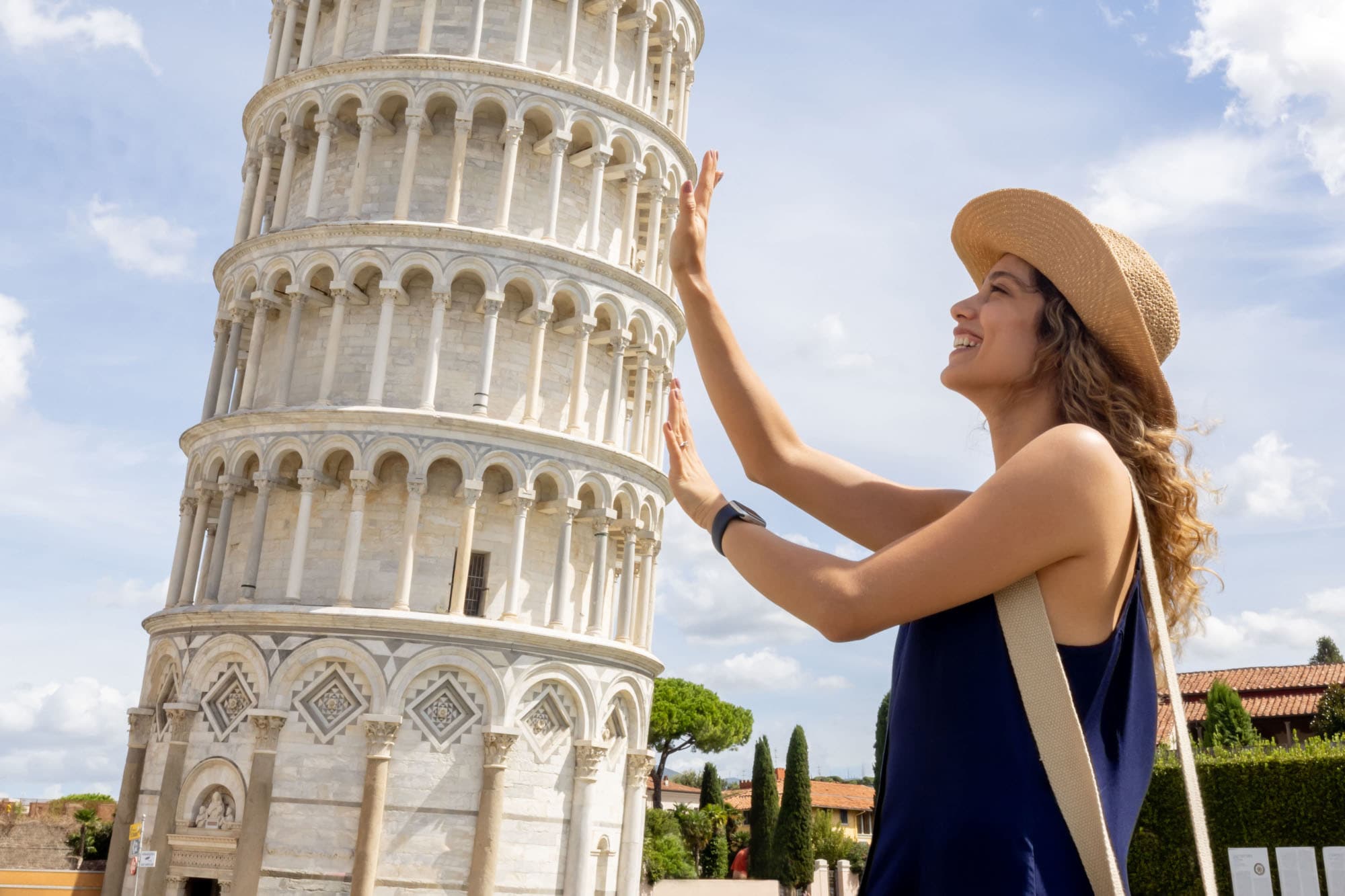 Profilansicht einer jungen Frau mit Strohhut, die aus der fotografischen Perspektive den Schiefen Turm von Pisa mit ihren Händen zu halten scheint. Profilansicht einer jungen Frau mit Strohhut, die aus der fotografischen Perspektive den Schiefen Turm von Pisa mit ihren Händen zu halten scheint.