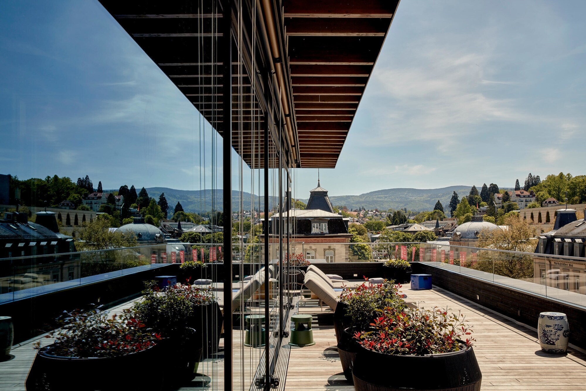 Große Balkonterrasse mit Liegestühlen vor grüner Hügellandschaft, die sich in der Glasfassade des Gebäudes spiegelt. Große Balkonterrasse mit Liegestühlen vor grüner Hügellandschaft, die sich in der Glasfassade des Gebäudes spiegelt.