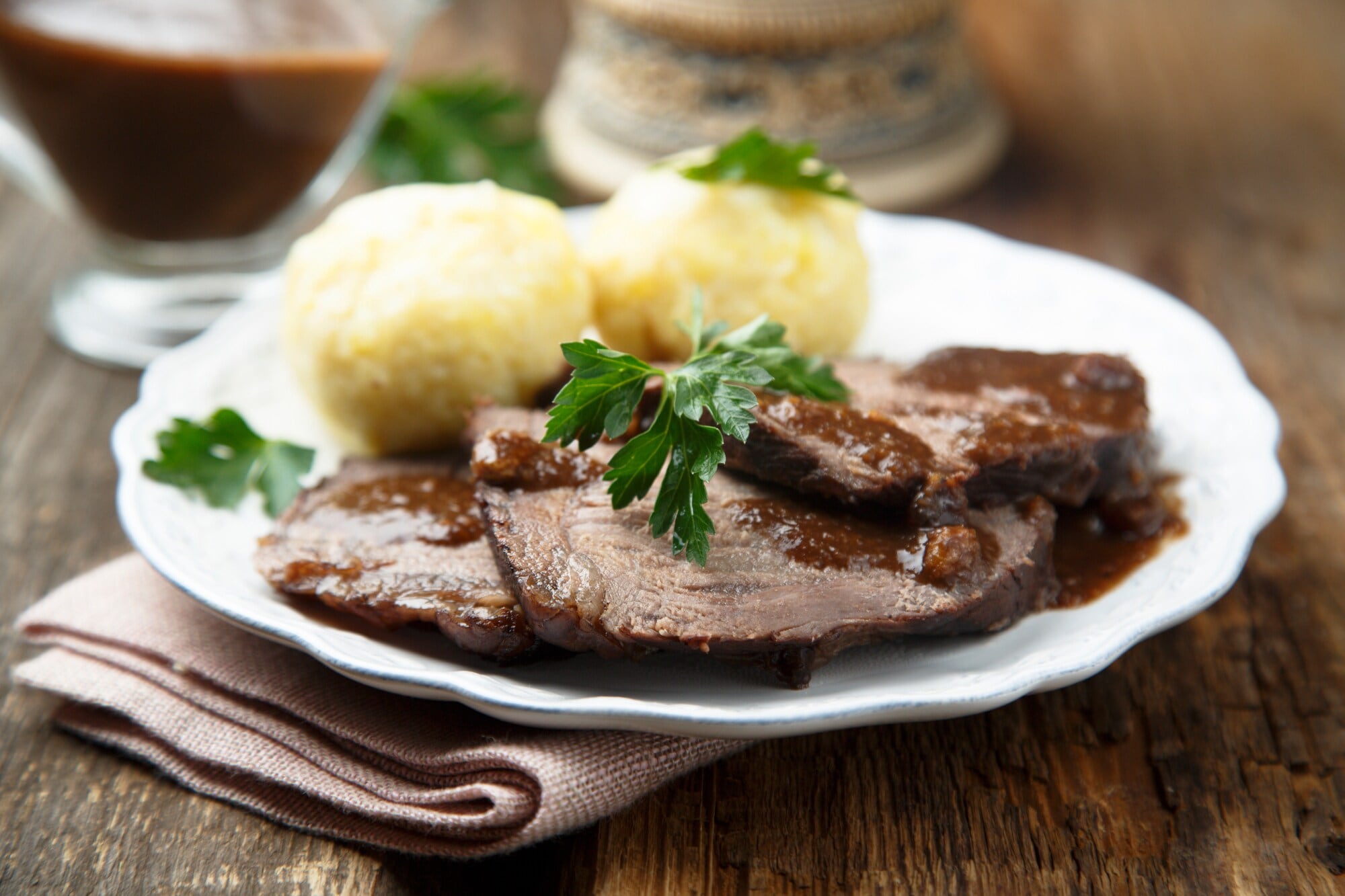 Sauerbraten mit brauner Soße an Klößen, angerichtet auf einem weißen Teller auf einem Holztisch. Sauerbraten mit brauner Soße an Klößen, angerichtet auf einem weißen Teller auf einem Holztisch.