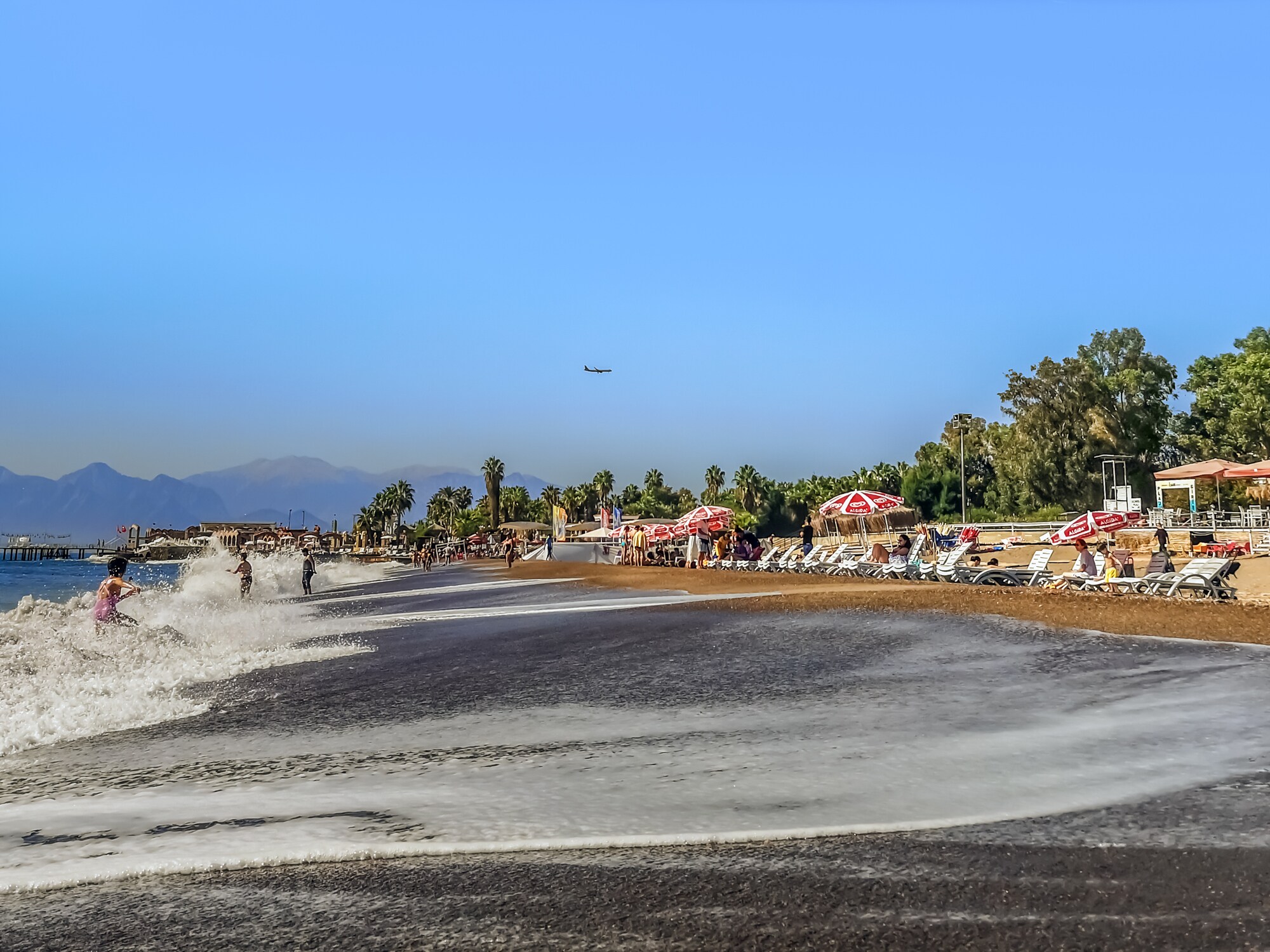 Kleiner Sandstrand mit Sonnenliegen und einigen Personen am Wasser, im Hintergrund Bäume und eine Gebirgskette.