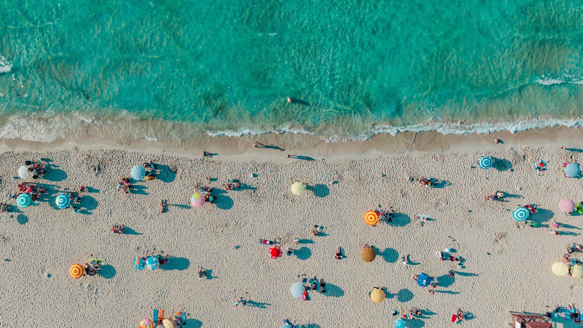 Belebter, weißer Sandstrand mit Sonnenschirmen am türkisblauen Wasser aus der Vogelperspektive.