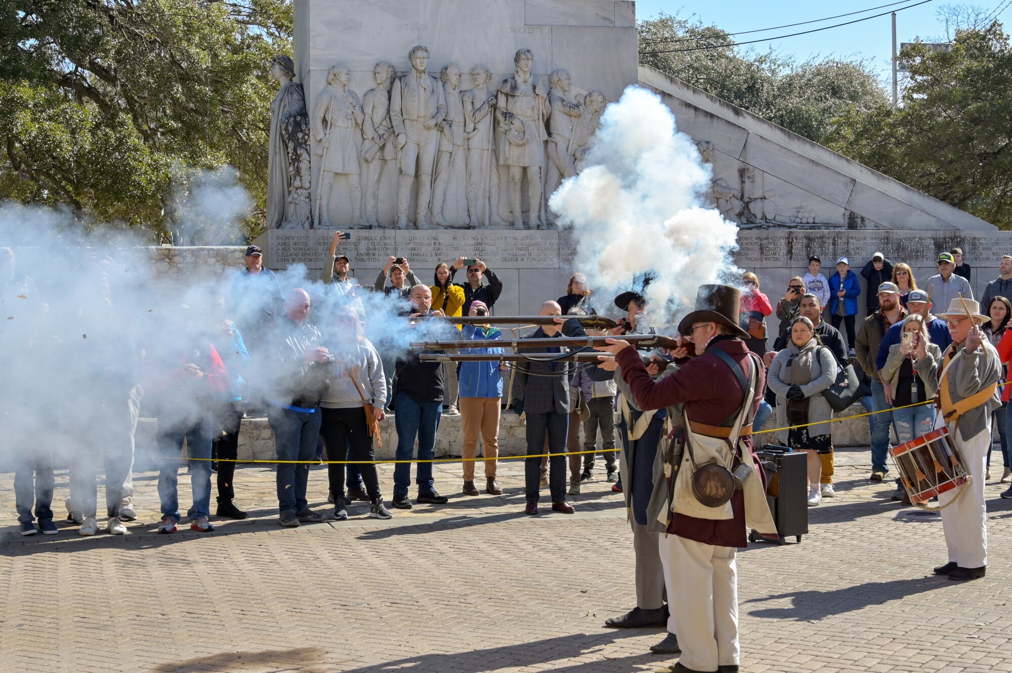 Eine Gruppe Männer in historischer Kleidung schießt bei einer Vorführung mit Langgewehren auf einem Platz, dahinter Zuschauende vor einem Denkmal.