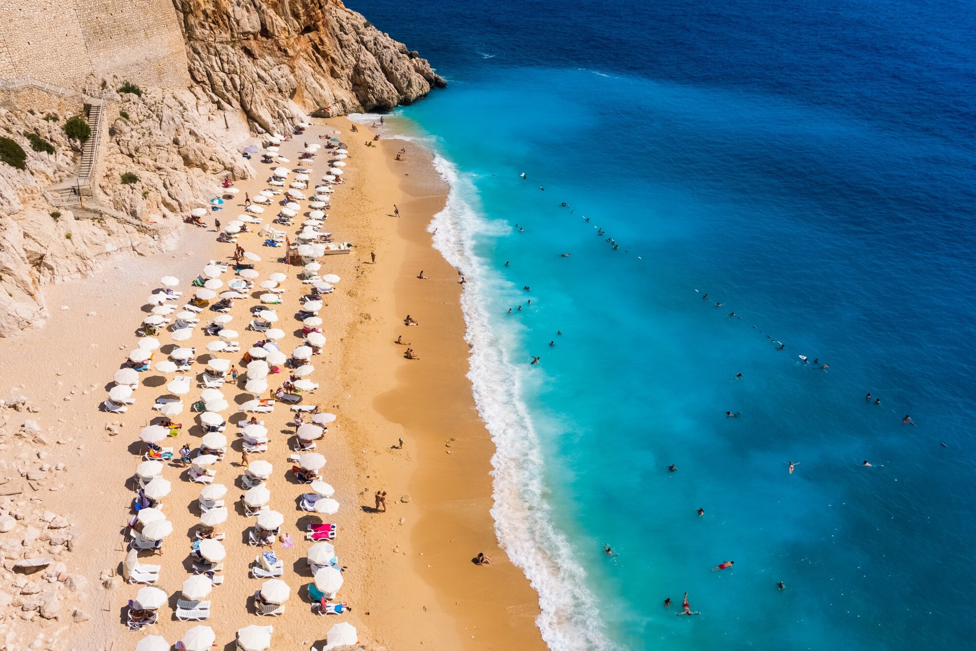 Luftaufnahme eines goldenen Sandstrandes mit Sonnenschirmen in einer Felsbucht mit Personen im türkisblauen Wasser. Luftaufnahme eines goldenen Sandstrandes mit Sonnenschirmen in einer Felsbucht mit Personen im türkisblauen Wasser.