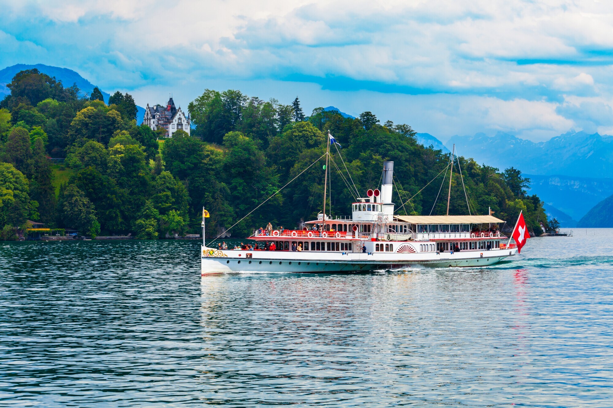 Ein Ausflugsschiff mit Schweizer Flagge auf einem von Wald umgebenen See vor Bergpanorama.