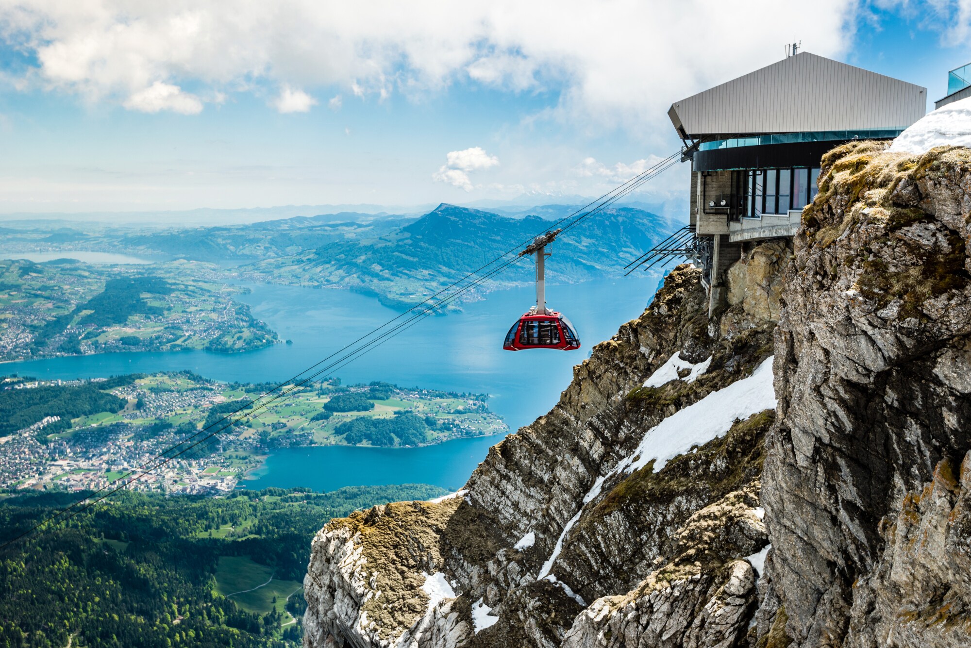 Eine rote Gondel einer Seilbahn an einem Berggipfel vor fjordartiger Landschaft des Vierwaldstättersees aus der Luft. Eine rote Gondel einer Seilbahn an einem Berggipfel vor fjordartiger Landschaft des Vierwaldstättersees aus der Luft.