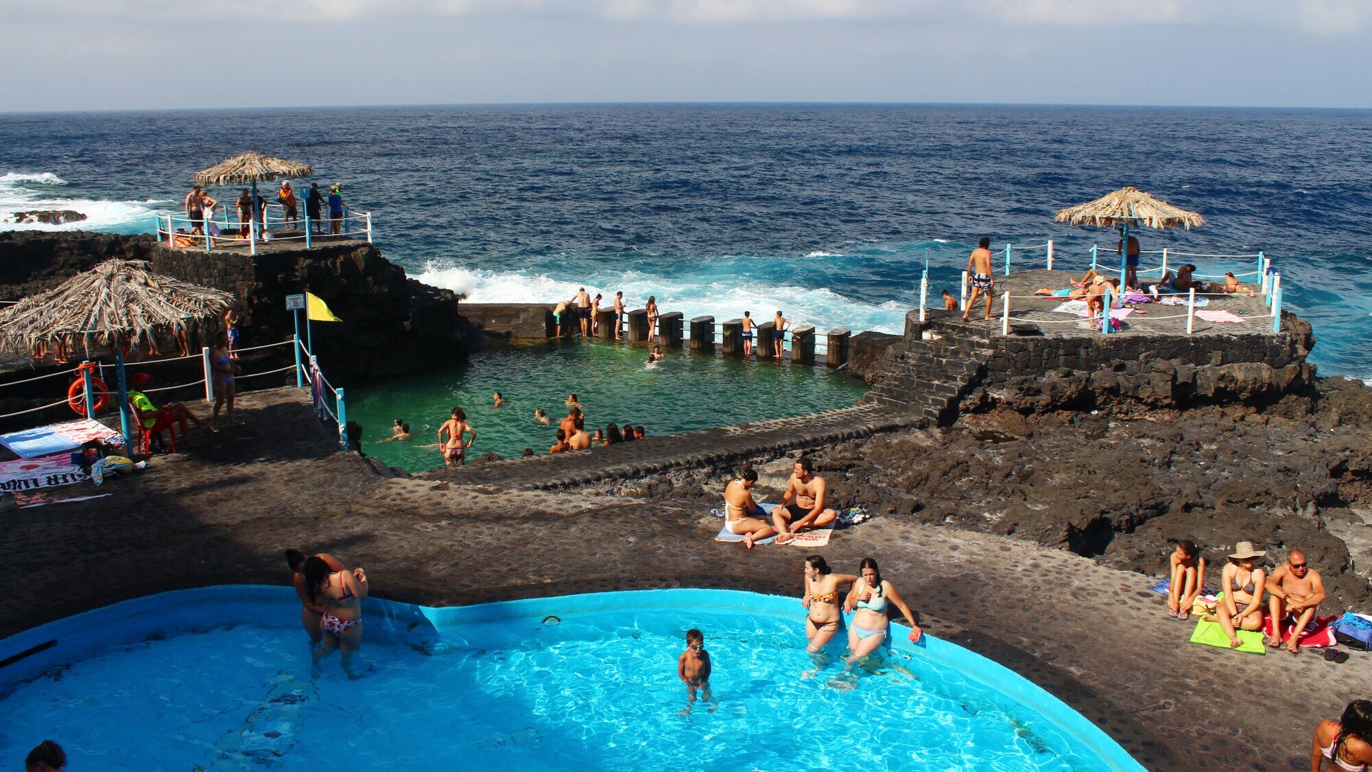Personen baden in Meerwasserbecken an einer Felsküste mit dunklem Vulkangestein. Personen baden in Meerwasserbecken an einer Felsküste mit dunklem Vulkangestein.