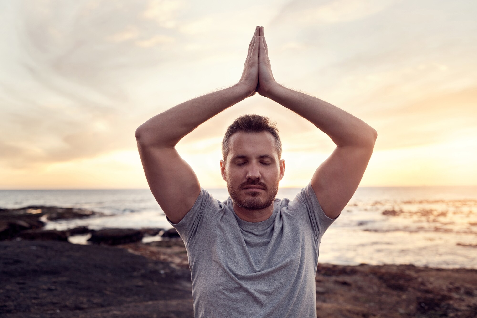 Ein Mann führt mit geschlossenen Augen eine Yogaübung mit über dem Kopf gefalteten Händen am Strand aus.