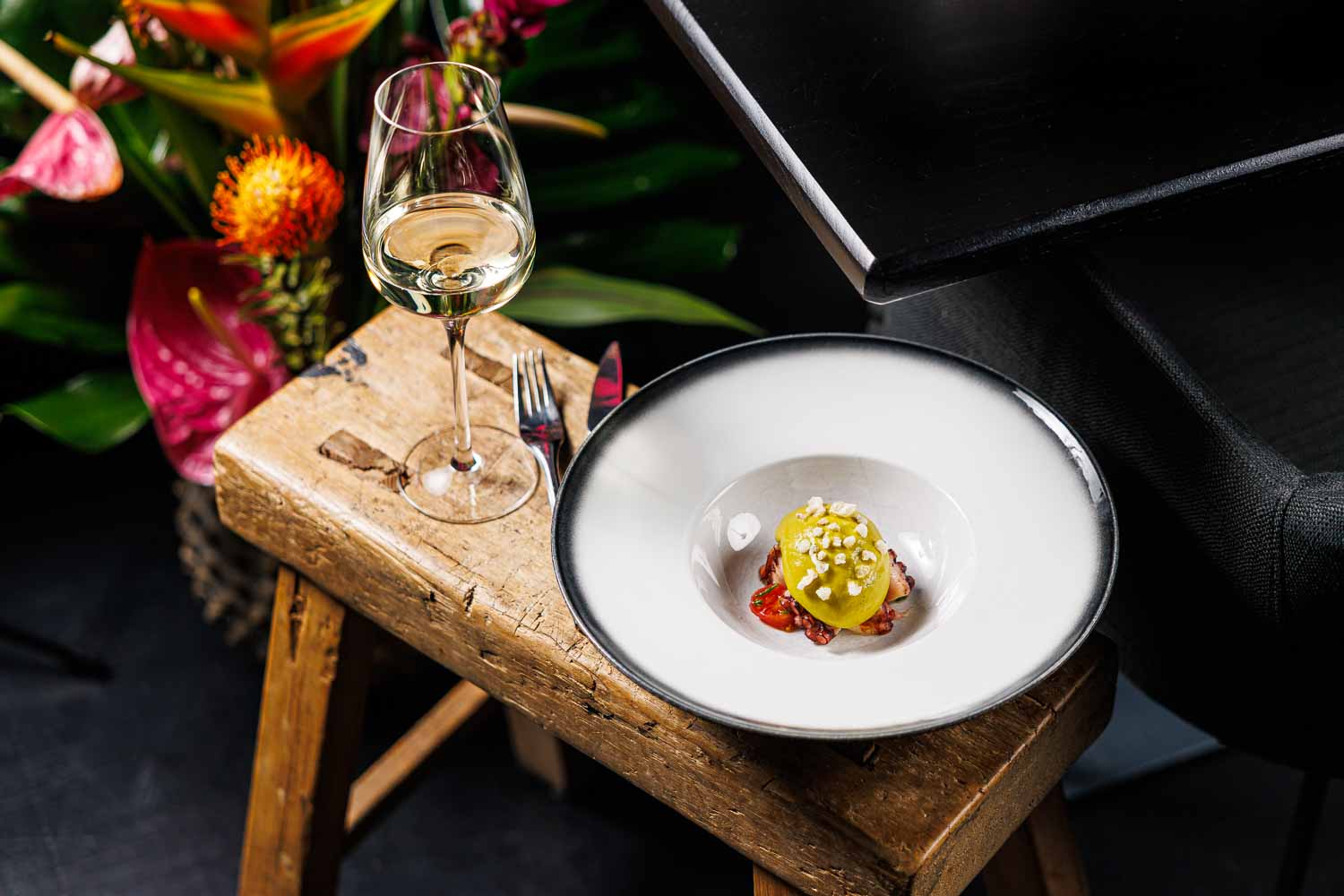 Ein weißer Teller mit einer kreativ angerichteten Vorspeise steht neben einem Glas Weißwein auf einem Holzschemel, daneben ein Blumenbouquet.