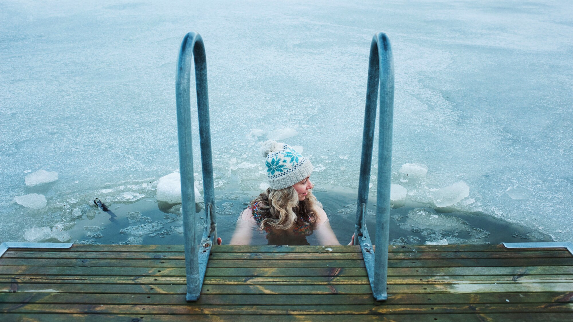 Eine Frau mit Wollmütze hält sich beim Baden in einem zugefrorenen See an dem Handlauf einer Treppe an einem Holzsteg fest.