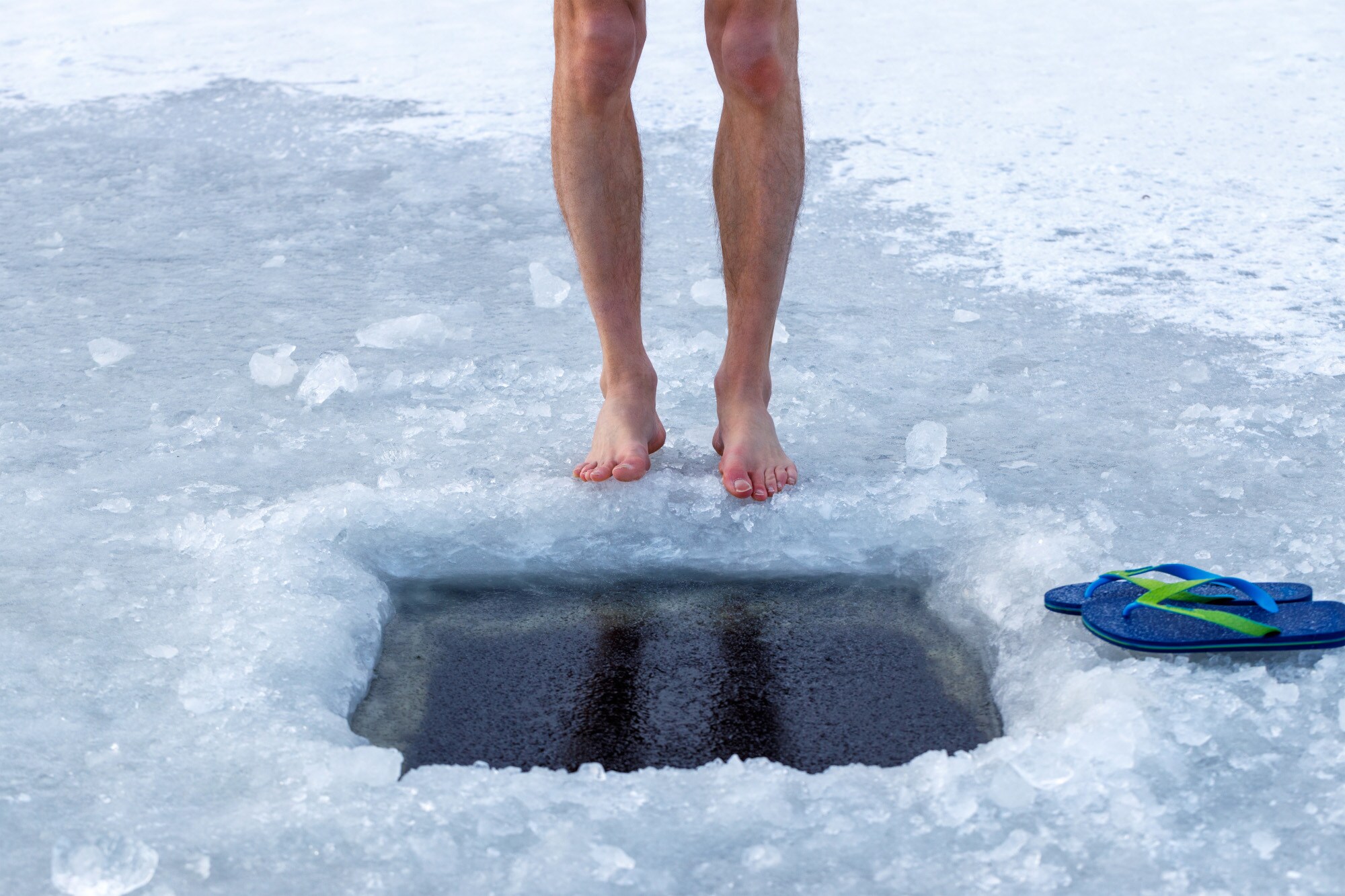 Zwei nackte Beine auf einem zugefrorenen See vor einem aufgebohrten Wasserloch, daneben zwei Badeschlappen.