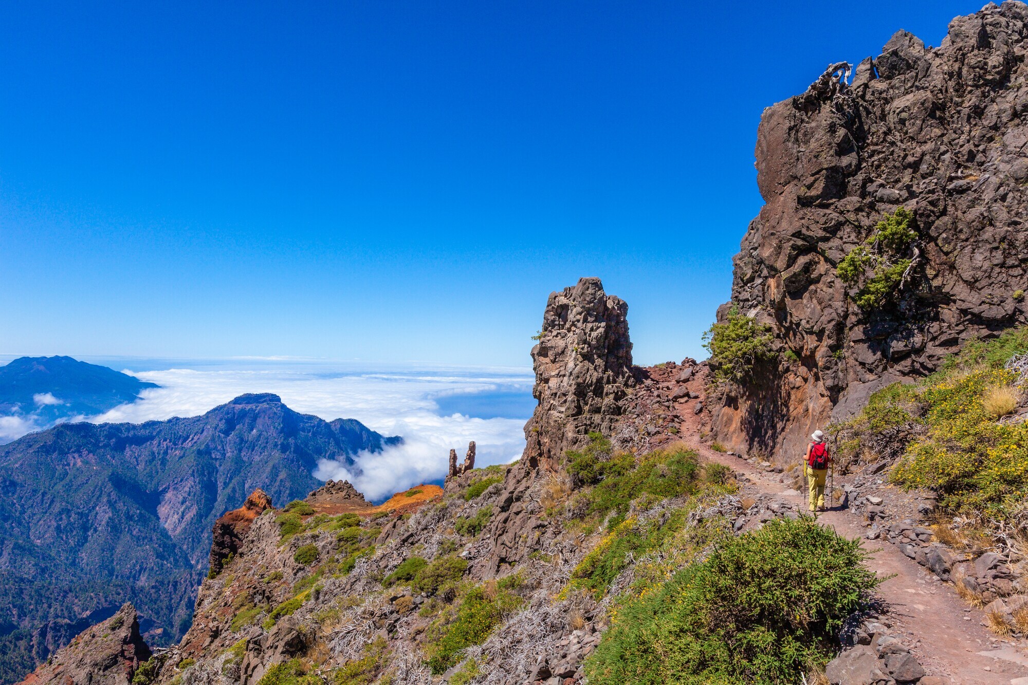 Eine Person auf einem Wanderweg auf einem Bergkamm mit rötlichen Felsformationen oberhalb der Wolkendecke unter blauem Himmel. Eine Person auf einem Wanderweg auf einem Bergkamm mit rötlichen Felsformationen oberhalb der Wolkendecke unter blauem Himmel.