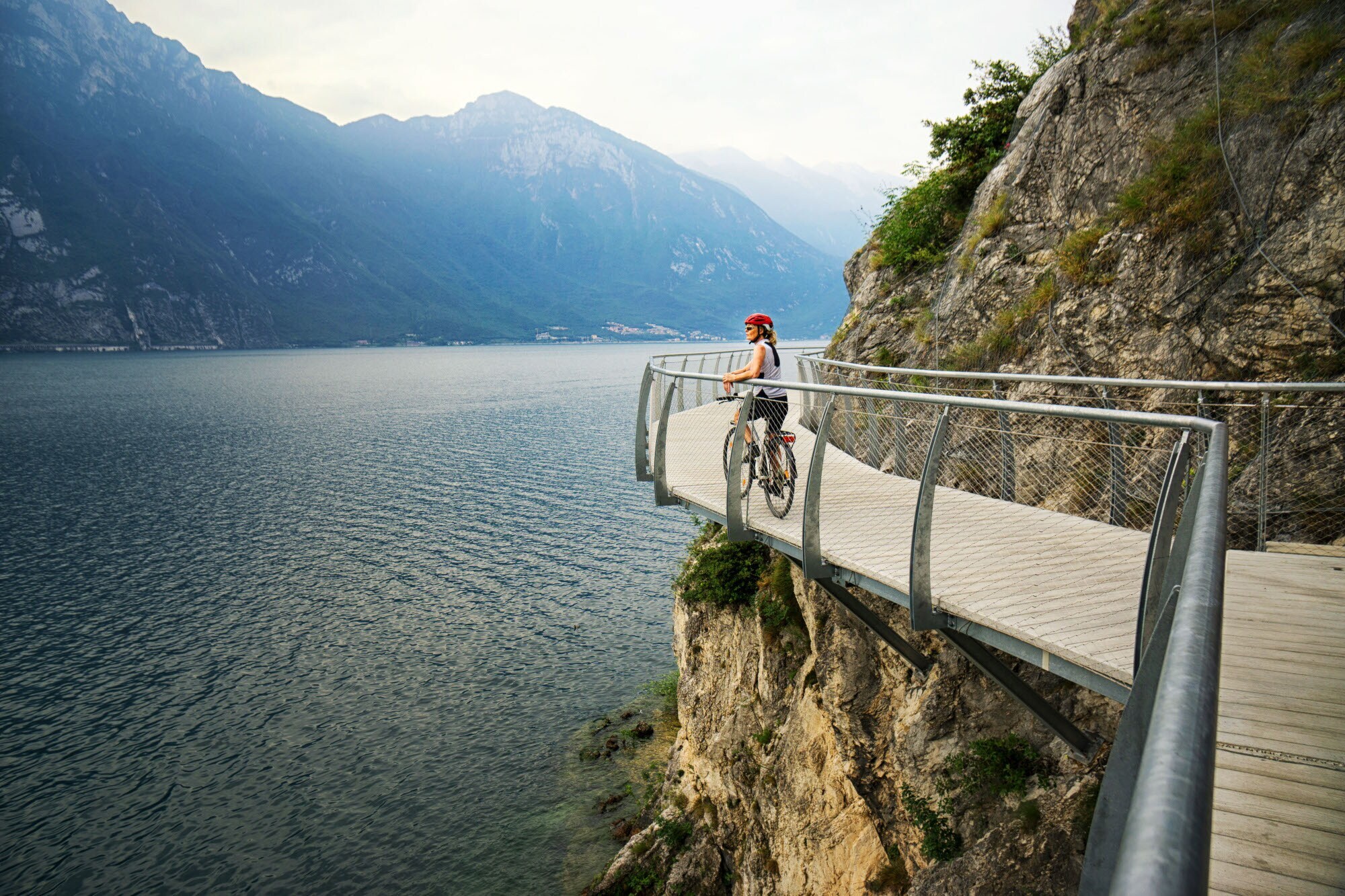 Eine Radfahrerin steht am Geländer eines Radweges an einem Felshang oberhalb eines Sees.