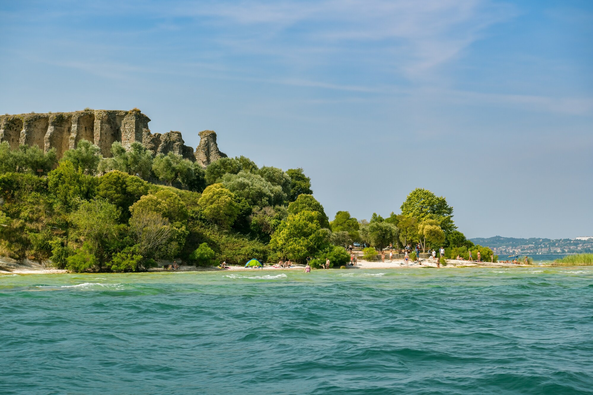 Eine antike Ruine, umgeben von üppiger Natur, an einem Sandstrand mit Personen am türkisblauen Wasser. Eine antike Ruine, umgeben von üppiger Natur, an einem Sandstrand mit Personen am türkisblauen Wasser.