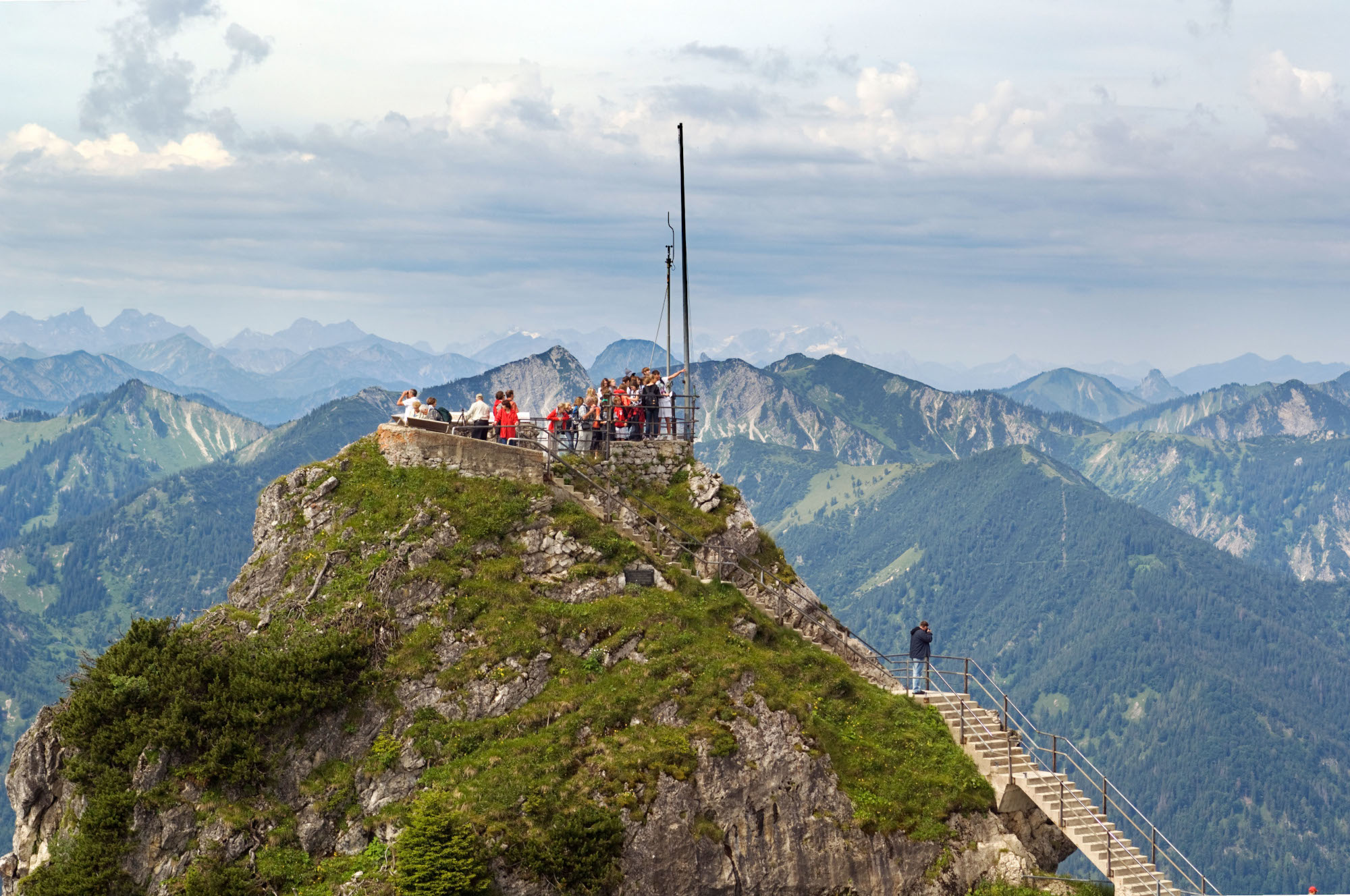 Personen auf einer Aussichtsplattform auf einem Berggipfel vor Alpenpanorama. Personen auf einer Aussichtsplattform auf einem Berggipfel vor Alpenpanorama.