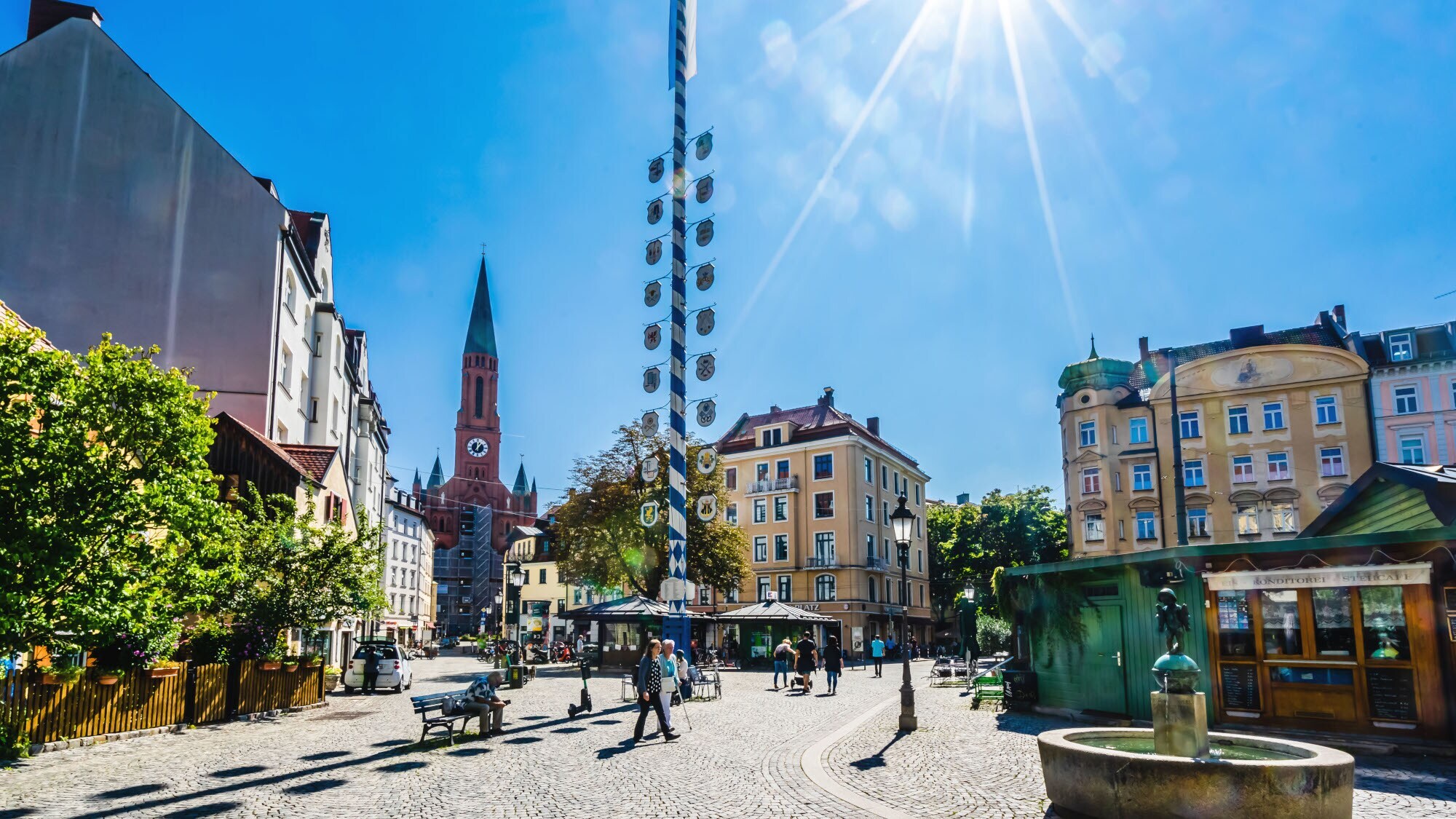 Ein Stadtteilplatz mit Kopfsteinpflaster und einem Maibaum in der Mitte bei strahlendem Sonnenschein. Ein Stadtteilplatz mit Kopfsteinpflaster und einem Maibaum in der Mitte bei strahlendem Sonnenschein.