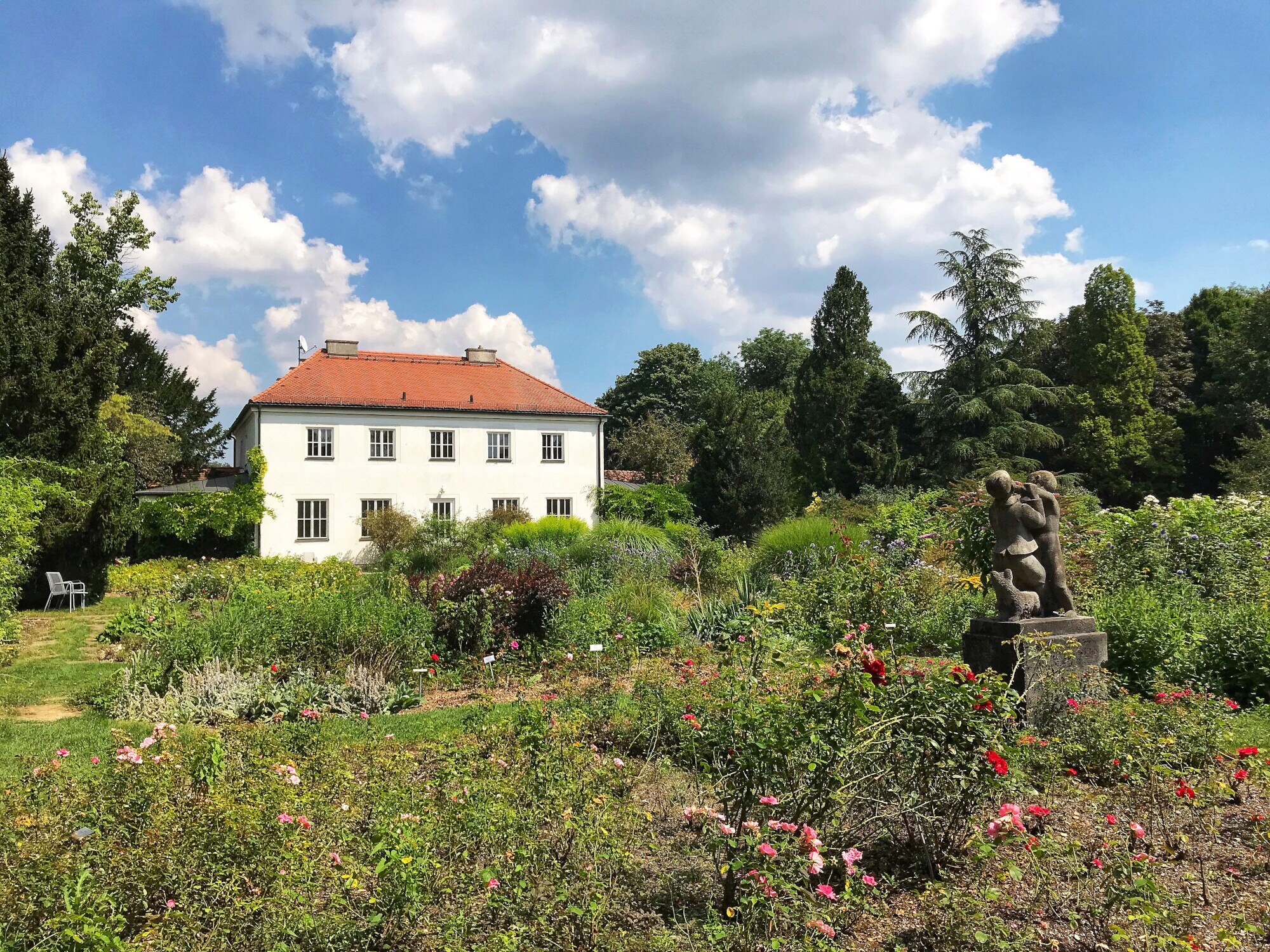 Blühende Rosenbüsche rund um eine kleine Statue in einem Rosengarten, im Hintergrund eine weiße Villa. Blühende Rosenbüsche rund um eine kleine Statue in einem Rosengarten, im Hintergrund eine weiße Villa.