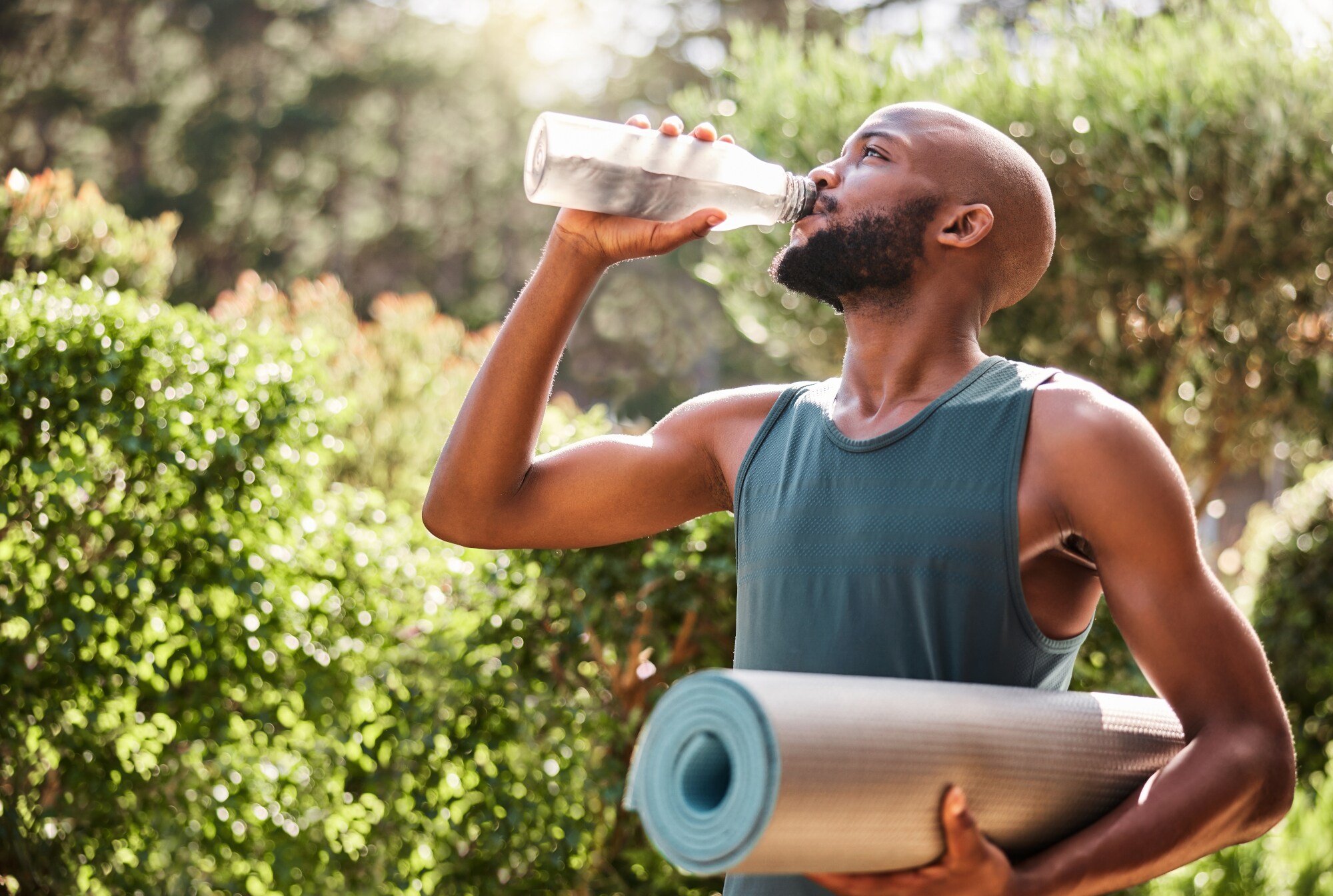 Ein junger, durchtrainierter Mann mit Yogamatte unter dem Arm trinkt aus einer Wasserflasche draußen in der Natur.