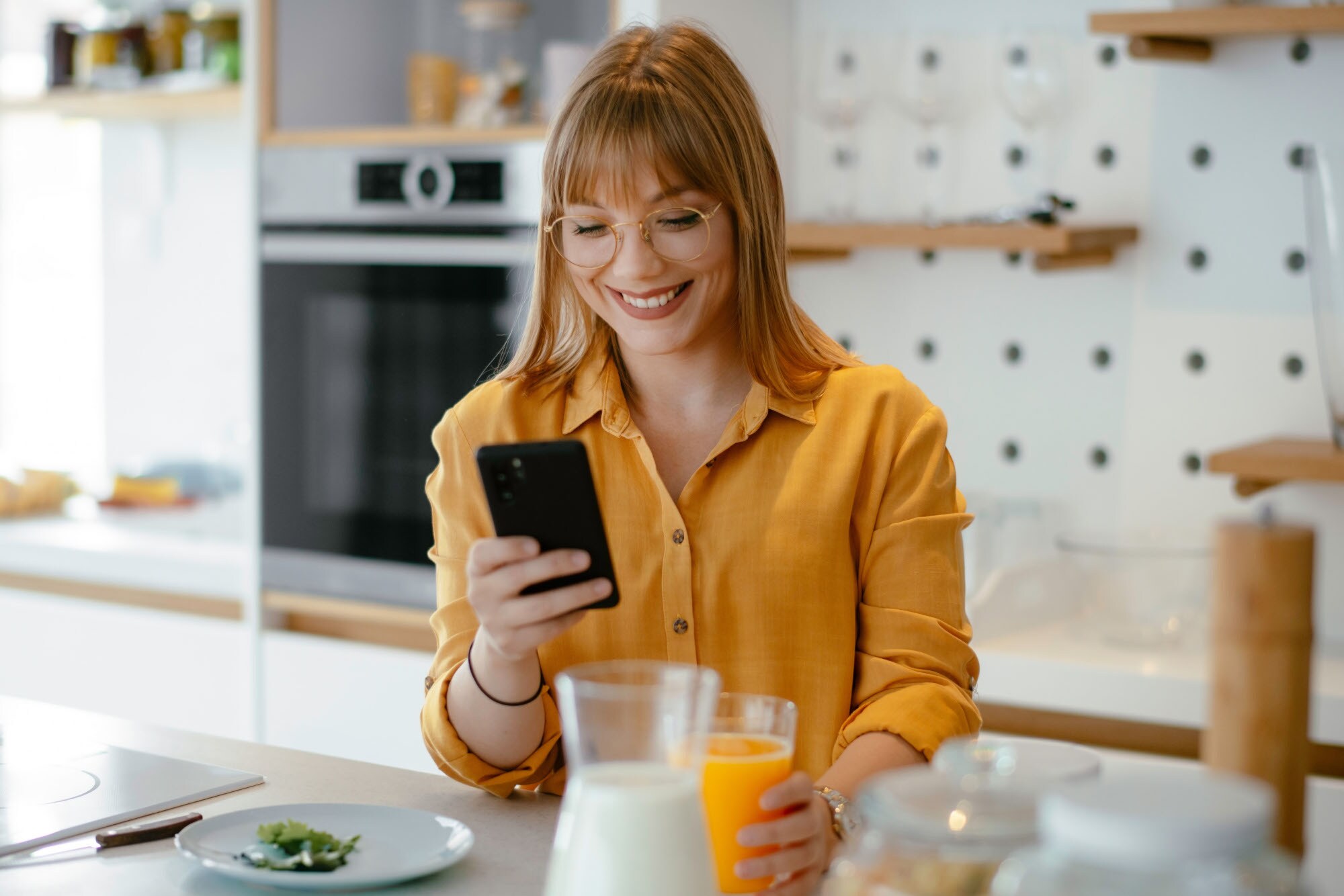Eine junge Frau in gelbem Hemd steht mit einem Glas Orangensaft in der Hand in der Küche und schaut lächelnd auf ihr Smartphone.