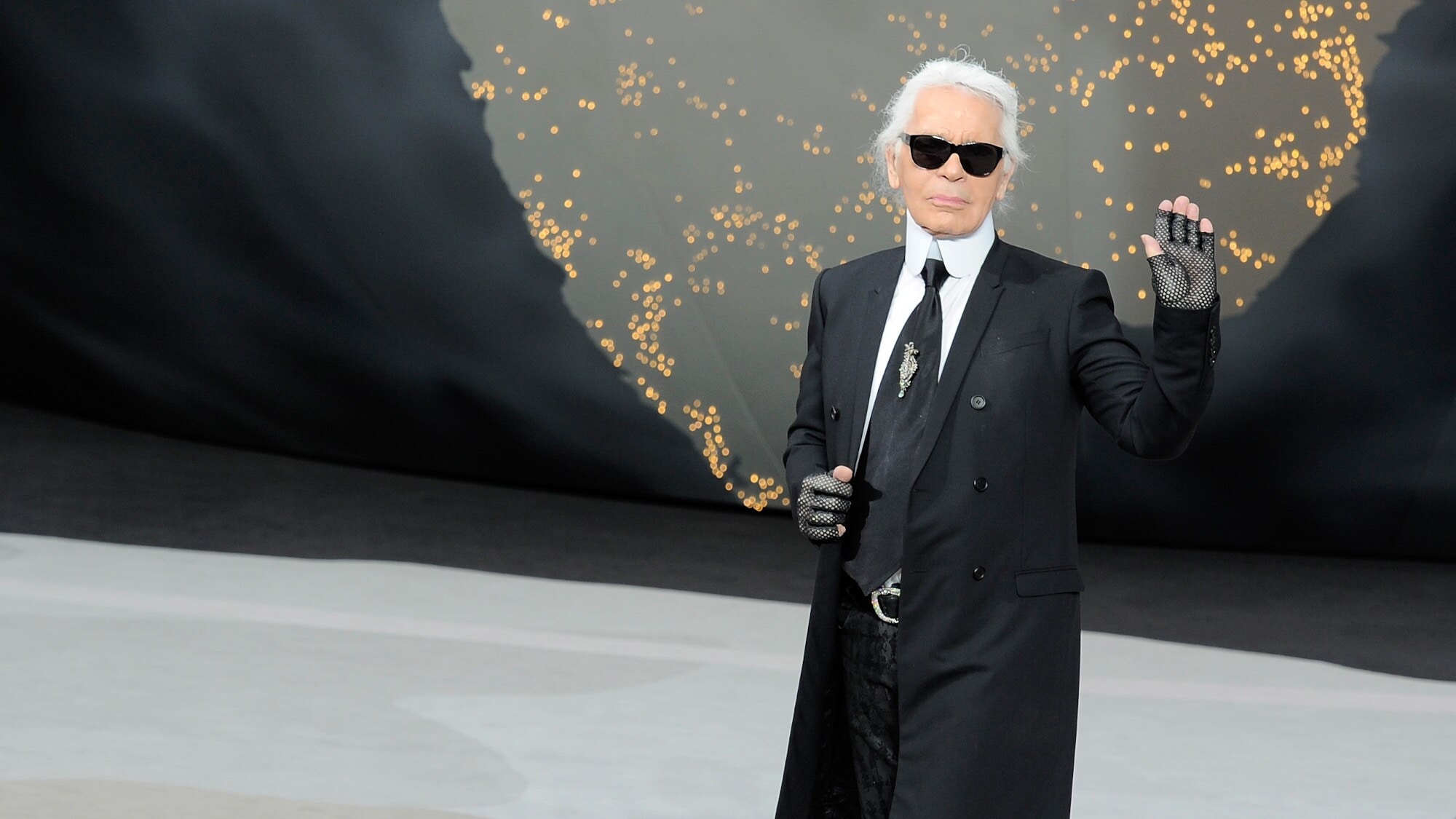 Karl Lagerfeld mit Sonnenbrille im schwarzen Jackett winkt vom Laufsteg einer Modenschau. Karl Lagerfeld mit Sonnenbrille im schwarzen Jackett winkt vom Laufsteg einer Modenschau.