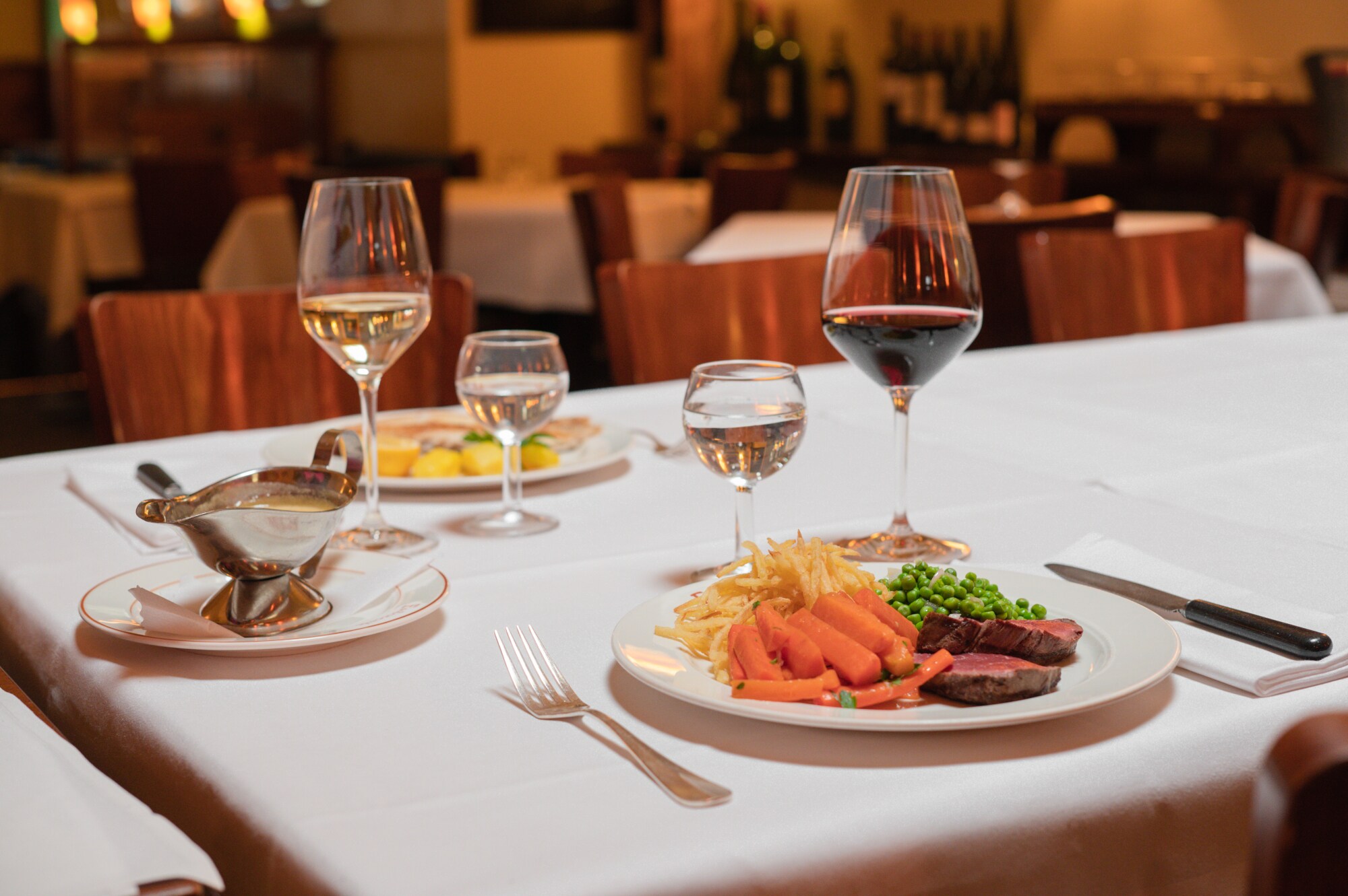 Zwei auf weißen Tellern angerichtete Speisen stehen neben Weingläsern auf einem Tisch mit weißer Stofftischdecke in einer Brasserie.