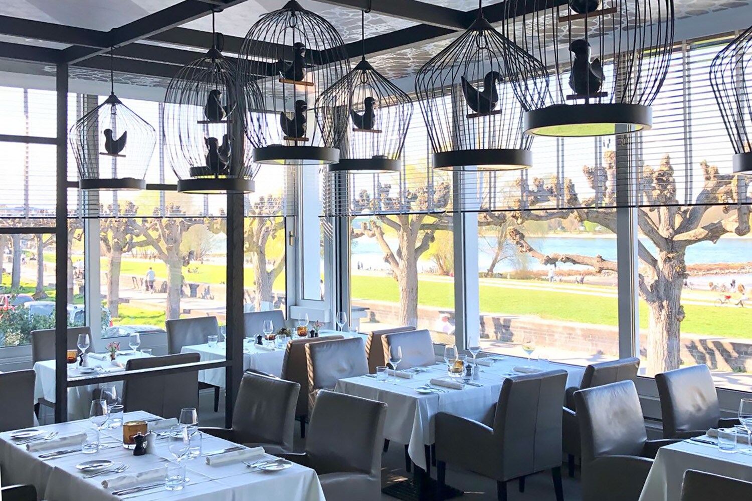 Speiseraum des Restaurants OX Royal mit eingedeckten Tischen und dekorativen Vogelkäfigen an der Decke in einem Wintergarten mit Panoramablick an der Uferpromenade eines Flusses.