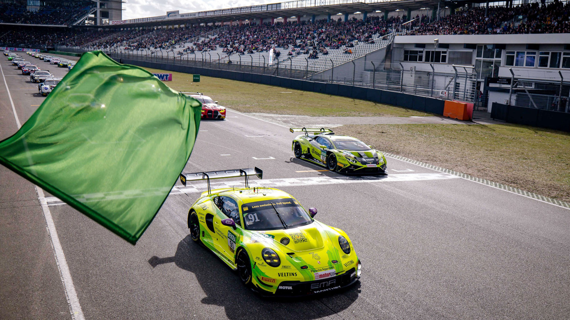 Ein Autorennen mit Zuschauern auf Tribünen, im Vordergrund eine grüne Flagge vor einem gelben Porsche.