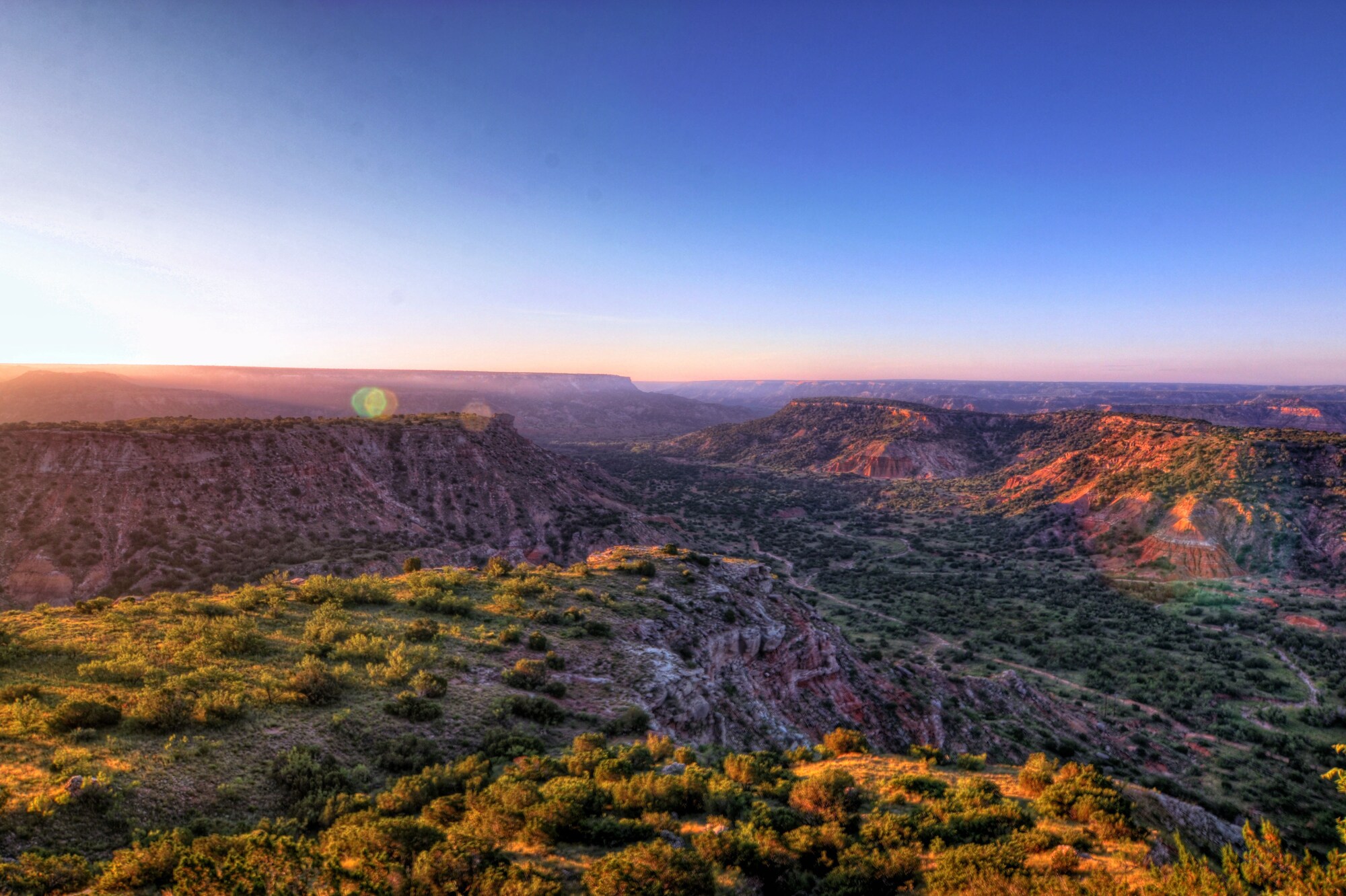 Landschaftspanorama eines weitläufigen Canyons mit roten Felsen und grüner Vegetation bei Sonnenuntergang.
