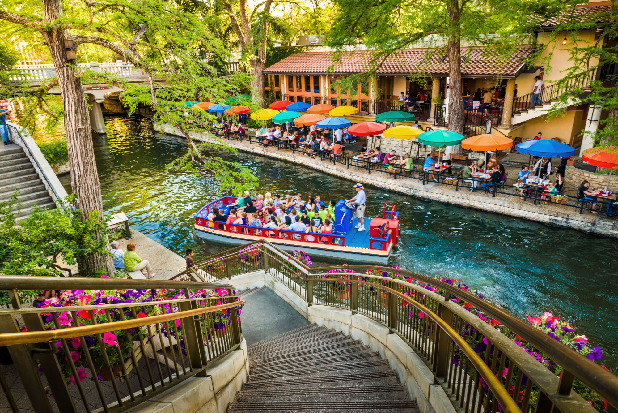 Ein Ausflugsboot fährt auf einem Wasserkanal an einem Restaurant mit Personen auf einer Terrasse mit bunten Sonnenschirmen vorbei, im Vordergrund eine mit Blumen dekorierte Treppe. Ein Ausflugsboot fährt auf einem Wasserkanal an einem Restaurant mit Personen auf einer Terrasse mit bunten Sonnenschirmen vorbei, im Vordergrund eine mit Blumen dekorierte Treppe.