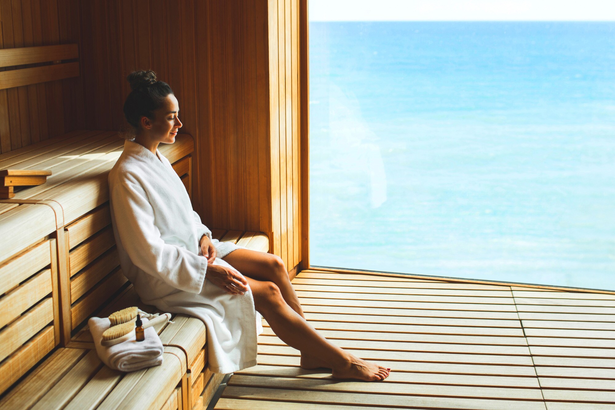 Eine junge Frau im weißen Bademantel sitzt entspannt in einer Holzsauna mit Panoramafenster am Meer. Eine junge Frau im weißen Bademantel sitzt entspannt in einer Holzsauna mit Panoramafenster am Meer.