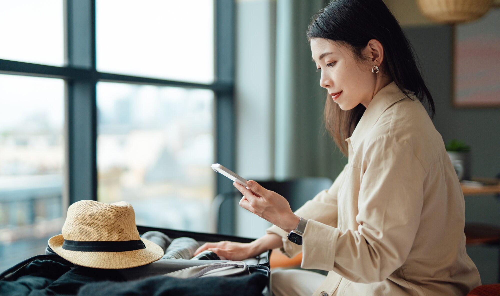 Eine Frau sitzt mit einem gepackten Koffer samt Strohhut auf einem Bett und schaut auf ihr Smartphone in der Hand. Eine Frau sitzt mit einem gepackten Koffer samt Strohhut auf einem Bett und schaut auf ihr Smartphone in der Hand.