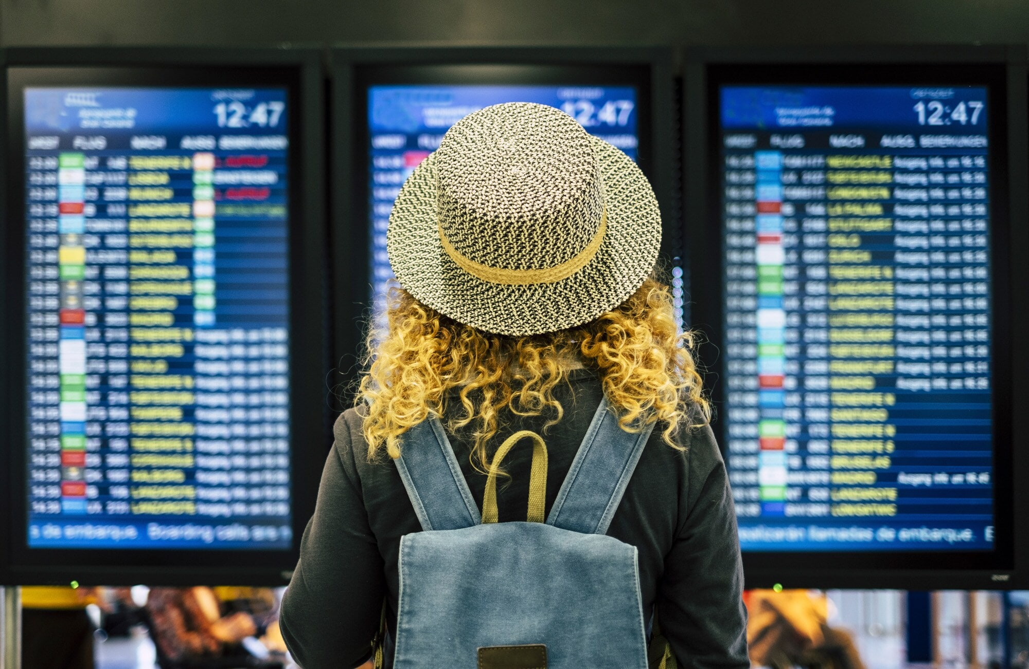 Rückansicht einer Frau mit Strohhut vor einer Anzeigetafel am Flughafen.