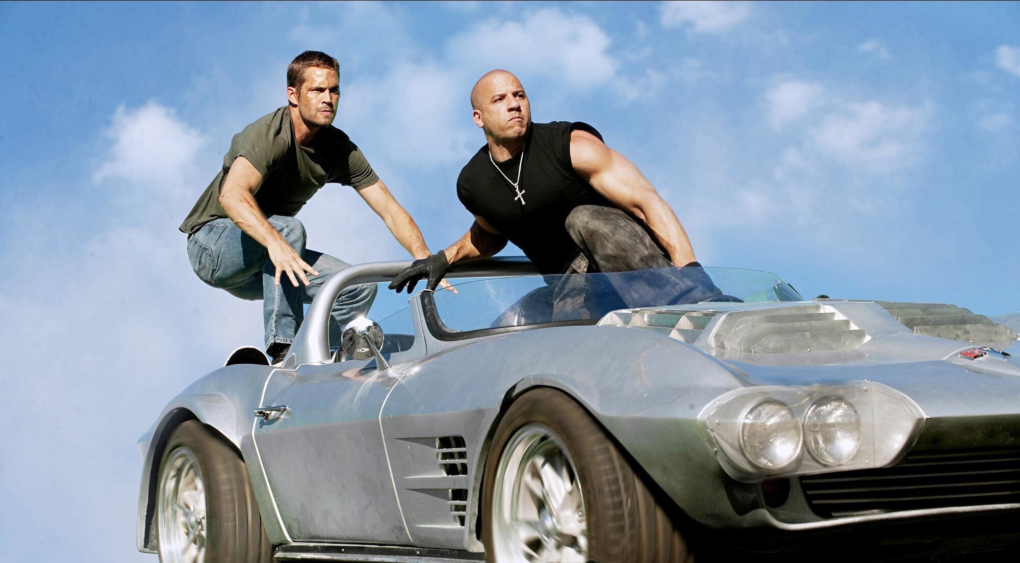 Paul Walker und Vin Diesel hocken auf einem silbernen Sportwagen ohne Verdeck.