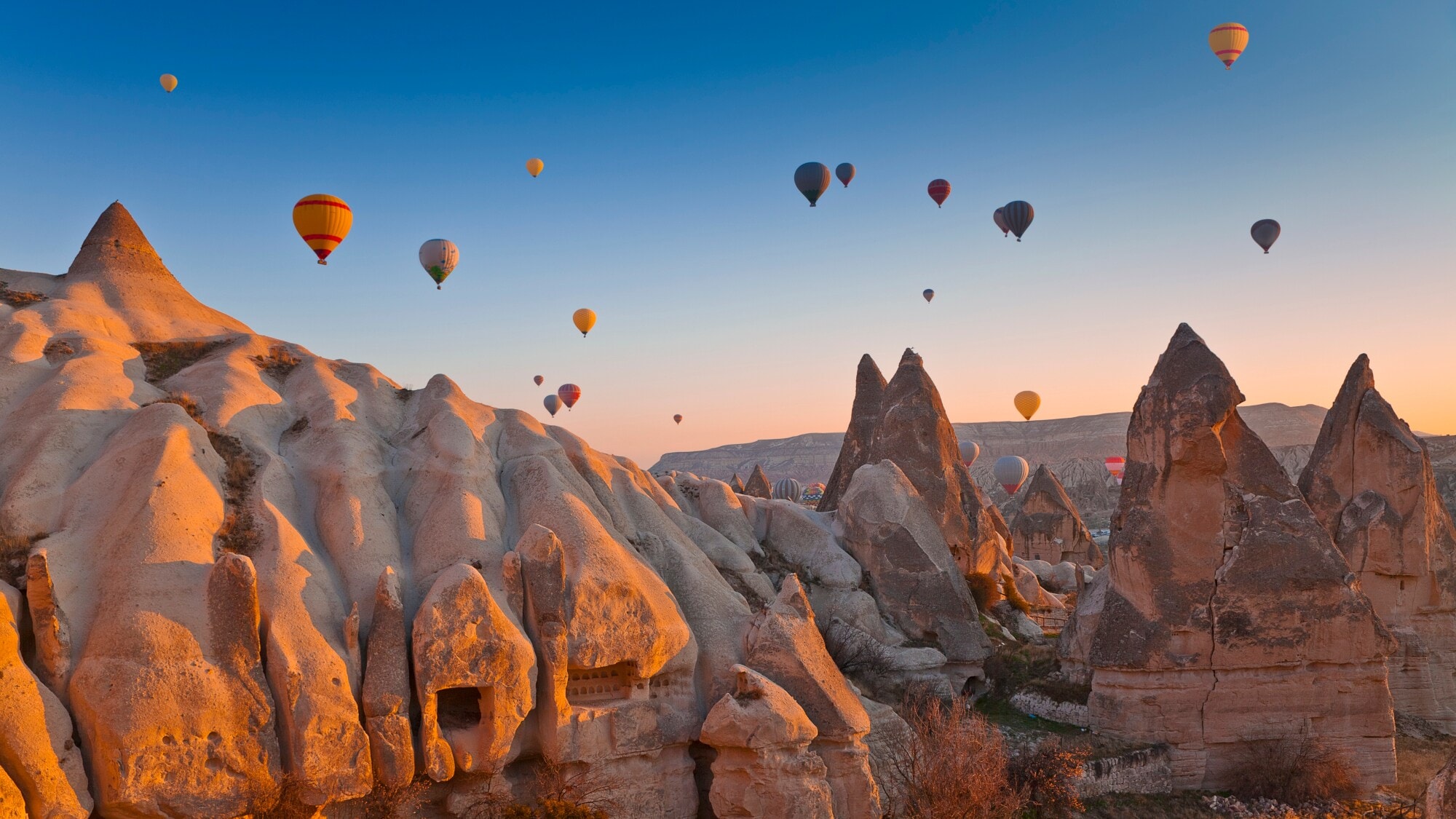 Felsige Landschaft in Kappadokien bei Sonnenuntergang mit zahlreichen schwebenden Heißluftballons im Hintergrund. Felsige Landschaft in Kappadokien bei Sonnenuntergang mit zahlreichen schwebenden Heißluftballons im Hintergrund.