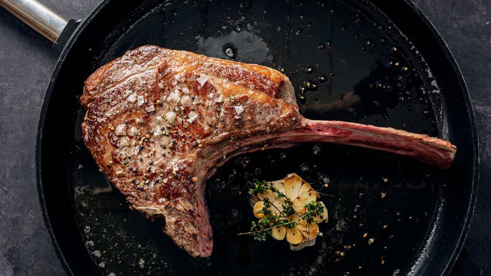 Ein scharf angebratenes Tomahawk-Steak in einer Pfanne. Daneben liegt eine aufgeschnittene Knoblauchknolle, die mit Thymian getoppt ist.