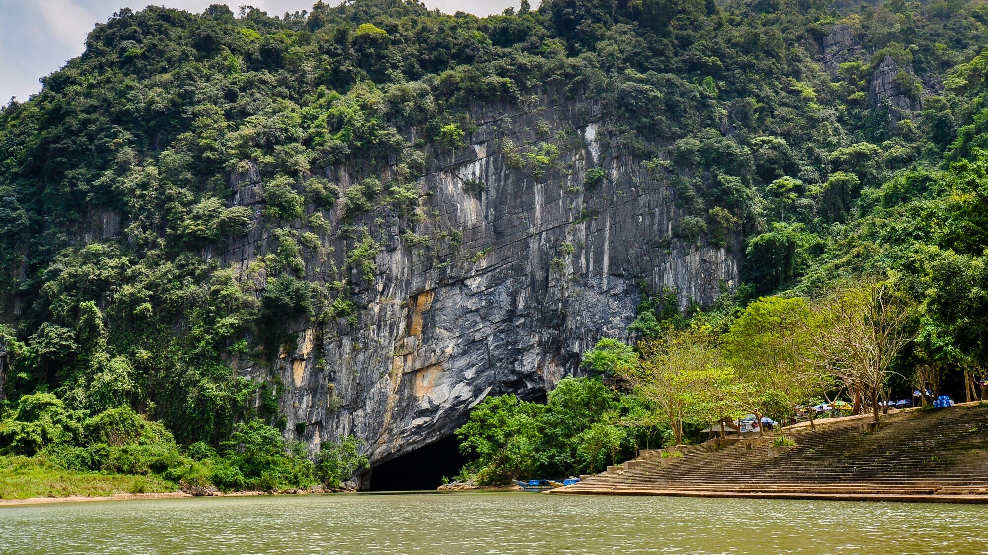 Eine Landschaft mit einem Fluss im Vordergrund und einer Felswand im Hintergrund, in die ein Höhleneingang führt.