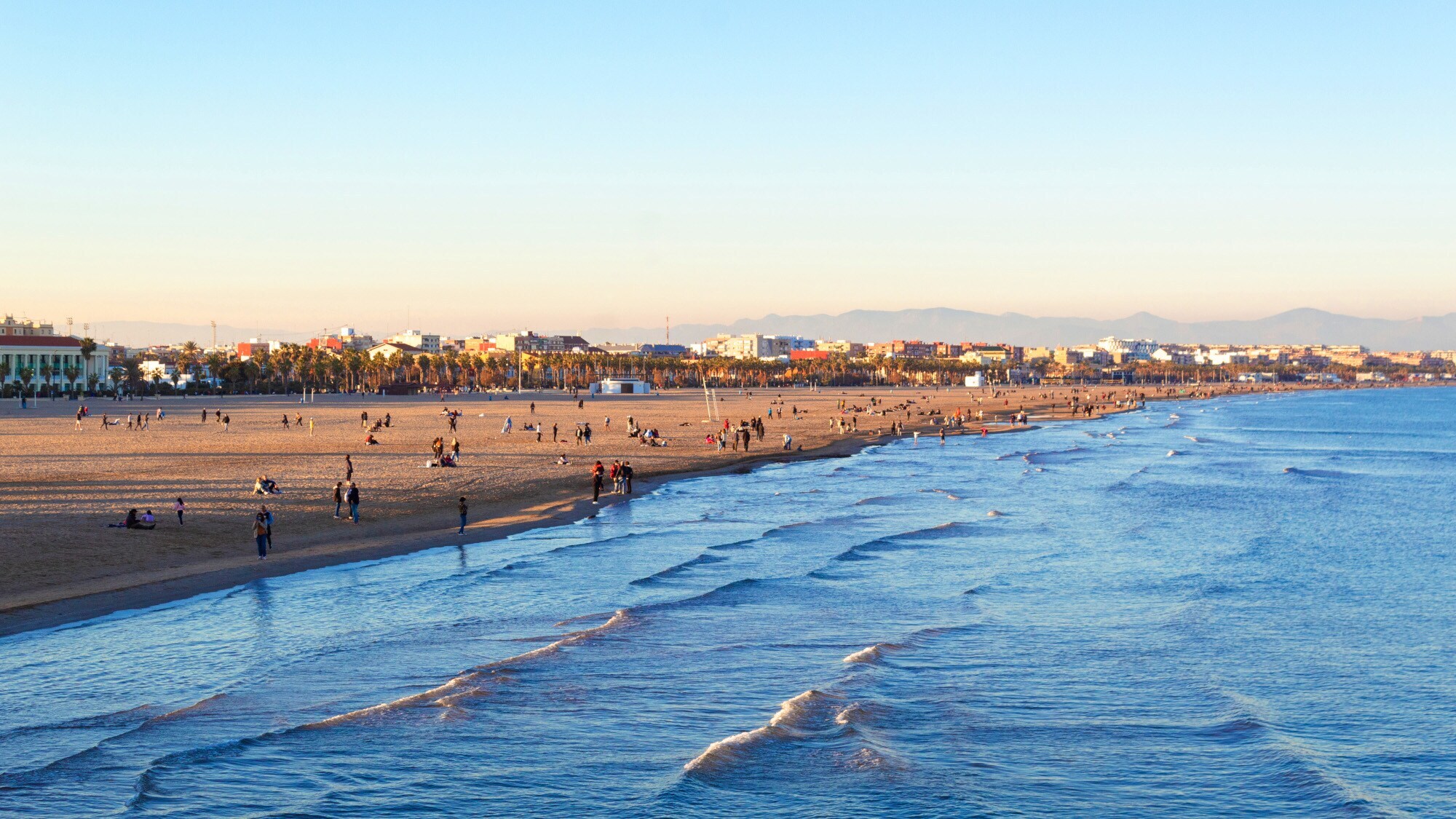 Personen an einem langen Sandstrand in urbaner Umgebung bei Sonnenuntergang. Personen an einem langen Sandstrand in urbaner Umgebung bei Sonnenuntergang.