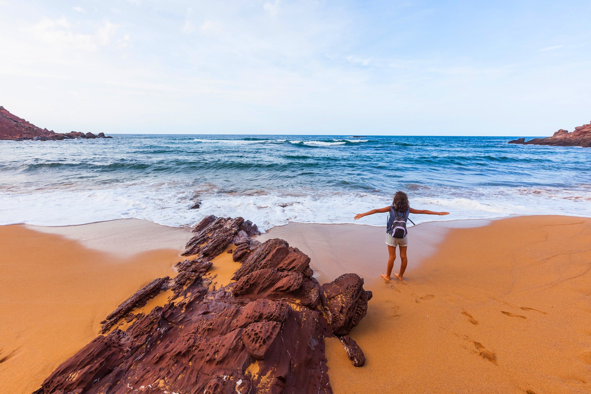 Eine Person steht mit dem Rücken zur Kamera und neben einem Felsen an einem Strand und schaut mit ausgebreiteten Armen aufs Meer